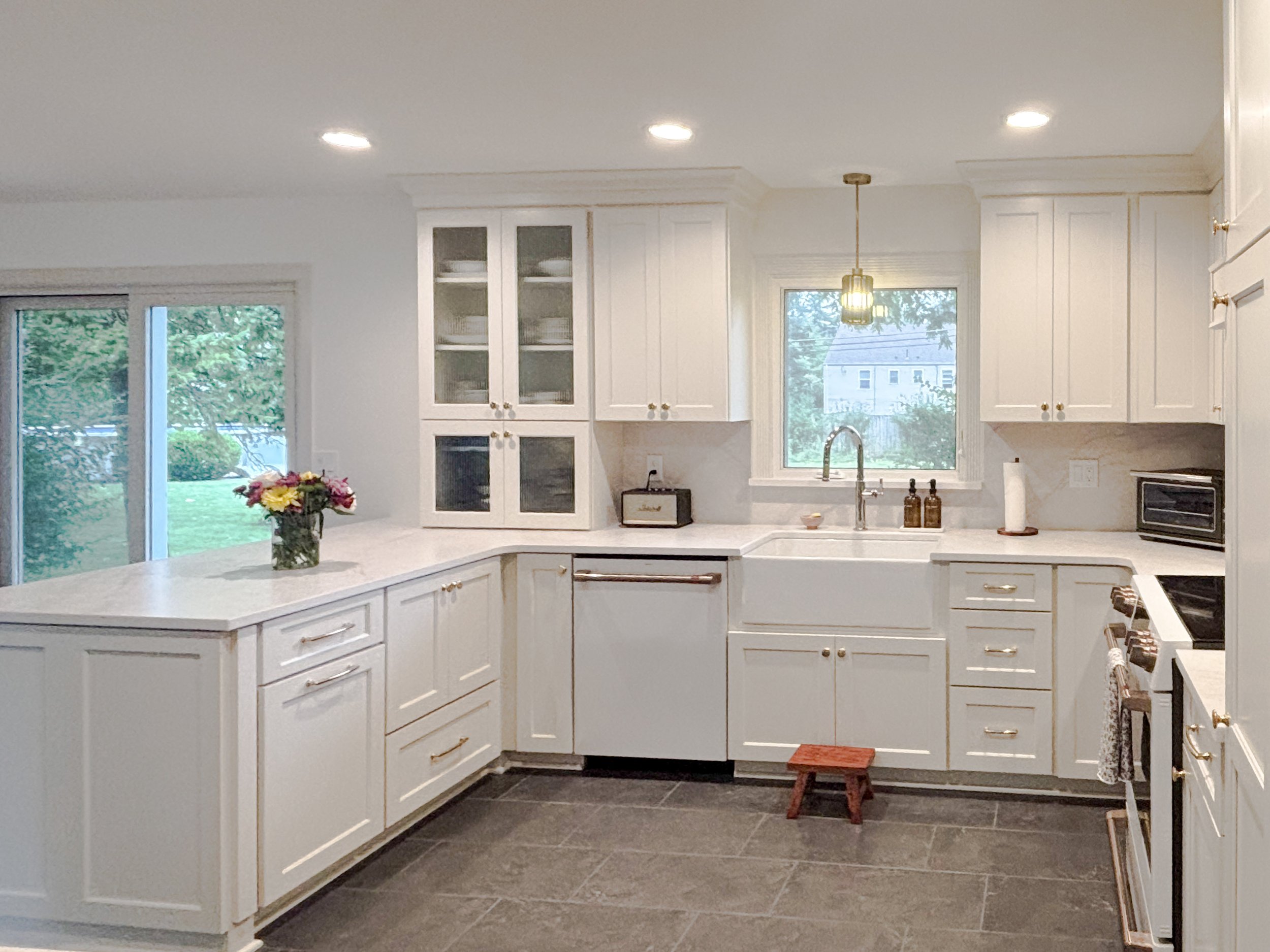 A kitchen with white cabinets, a marble countertop, a sink under a window, and a sliding glass door leading outside. There are two pendant lights, a small wooden stool, flowers on the counter, and a coffee maker.