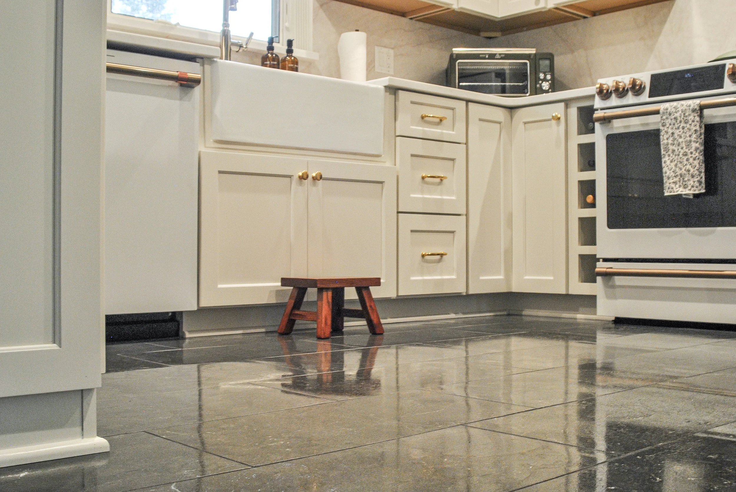Kitchen with white cabinets, black countertop, and a black oven. A small wooden stool is visible on the black tiled floor. There are two amber glass bottles and a paper towel roll on the counter near the window.