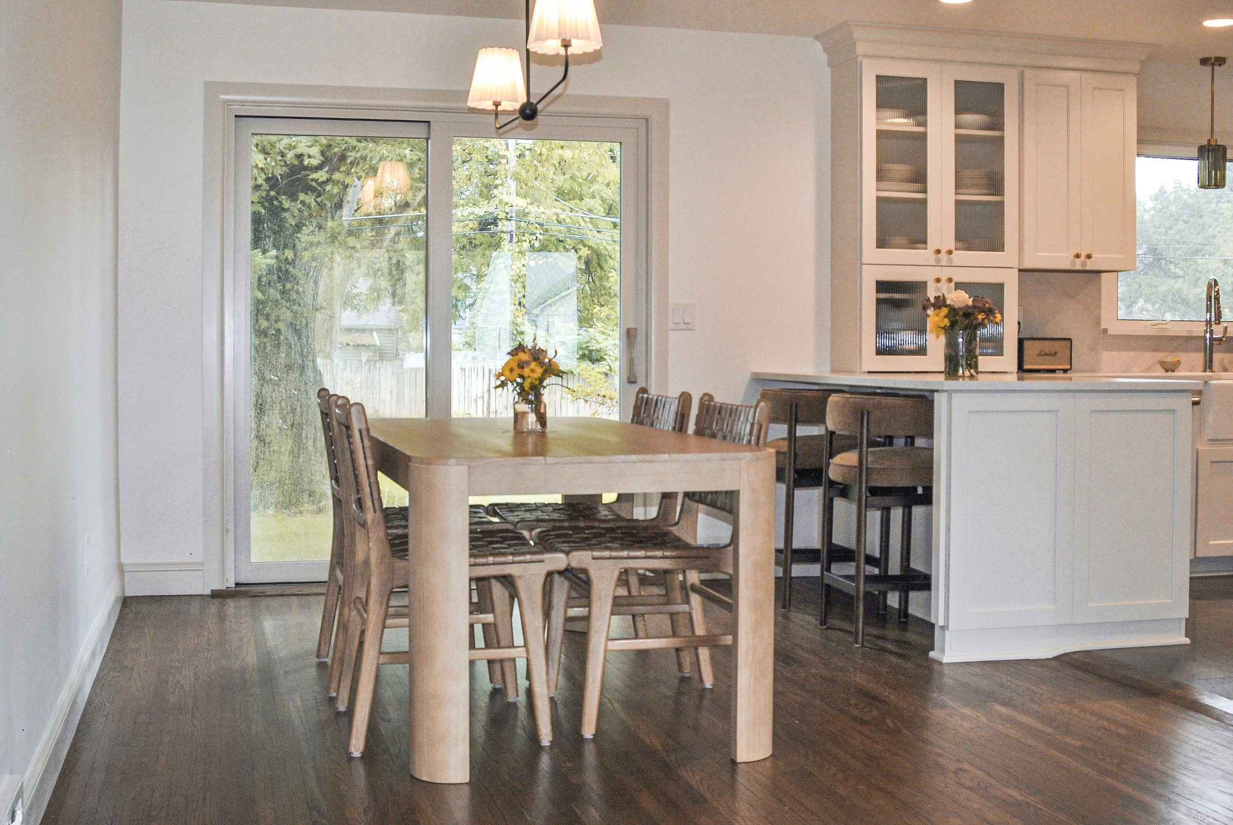 A dining area with a wooden table and chairs next to a large sliding glass door, with a view of a backyard with trees. There are flower vases with yellow and purple flowers on the table, and a kitchen area with white cabinets and a kitchen sink visible in the background. The room has hardwood flooring and hanging ceiling lights.