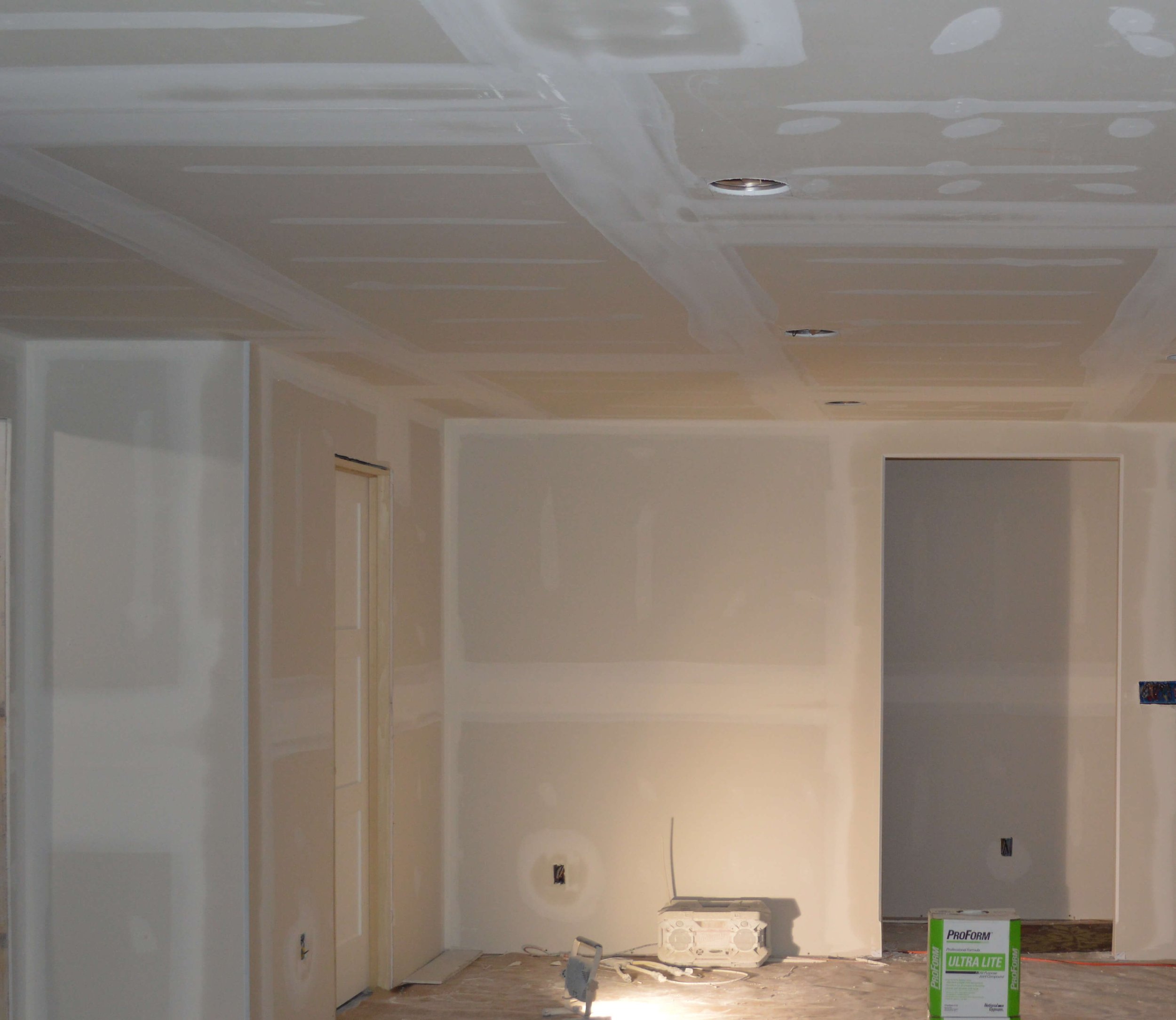 A room under construction or renovation with drywall installed on the ceiling and walls. There are holes cut in the ceiling for recessed lighting. Some construction materials and tools are on the floor, including a box of ProForm Ultra Lite joint compound.