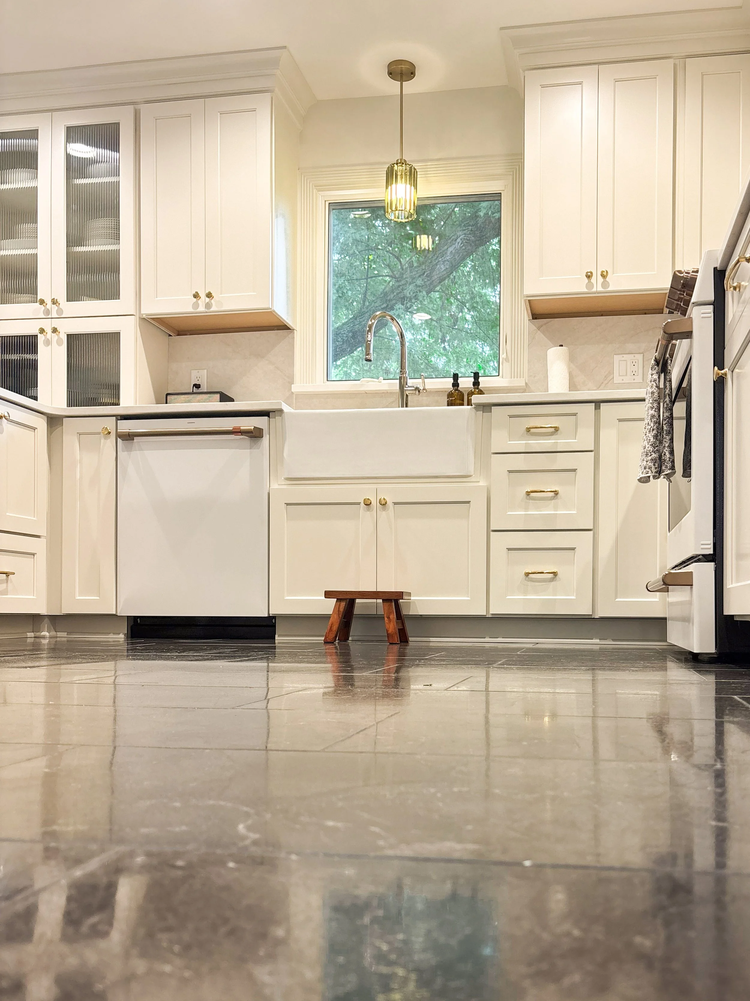 A kitchen with white cabinets, a large window above the sink showing green trees outside, and a gold pendant light hanging from the ceiling. The floor is made of shiny tiles and there is a small wooden stool in front of the sink.