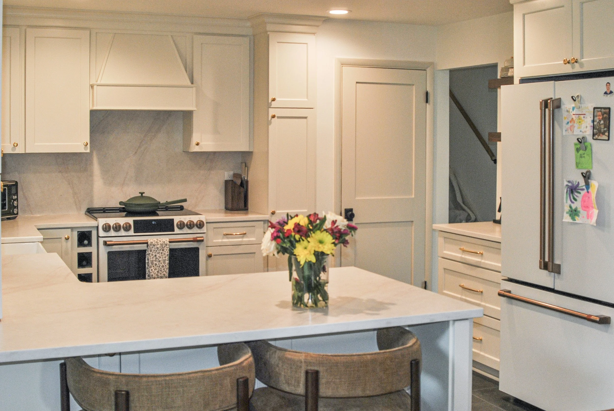A modern kitchen with white cabinets, a stove with a pot on top, a vase of flowers on a central island, and a refrigerator with colorful magnets and photos.