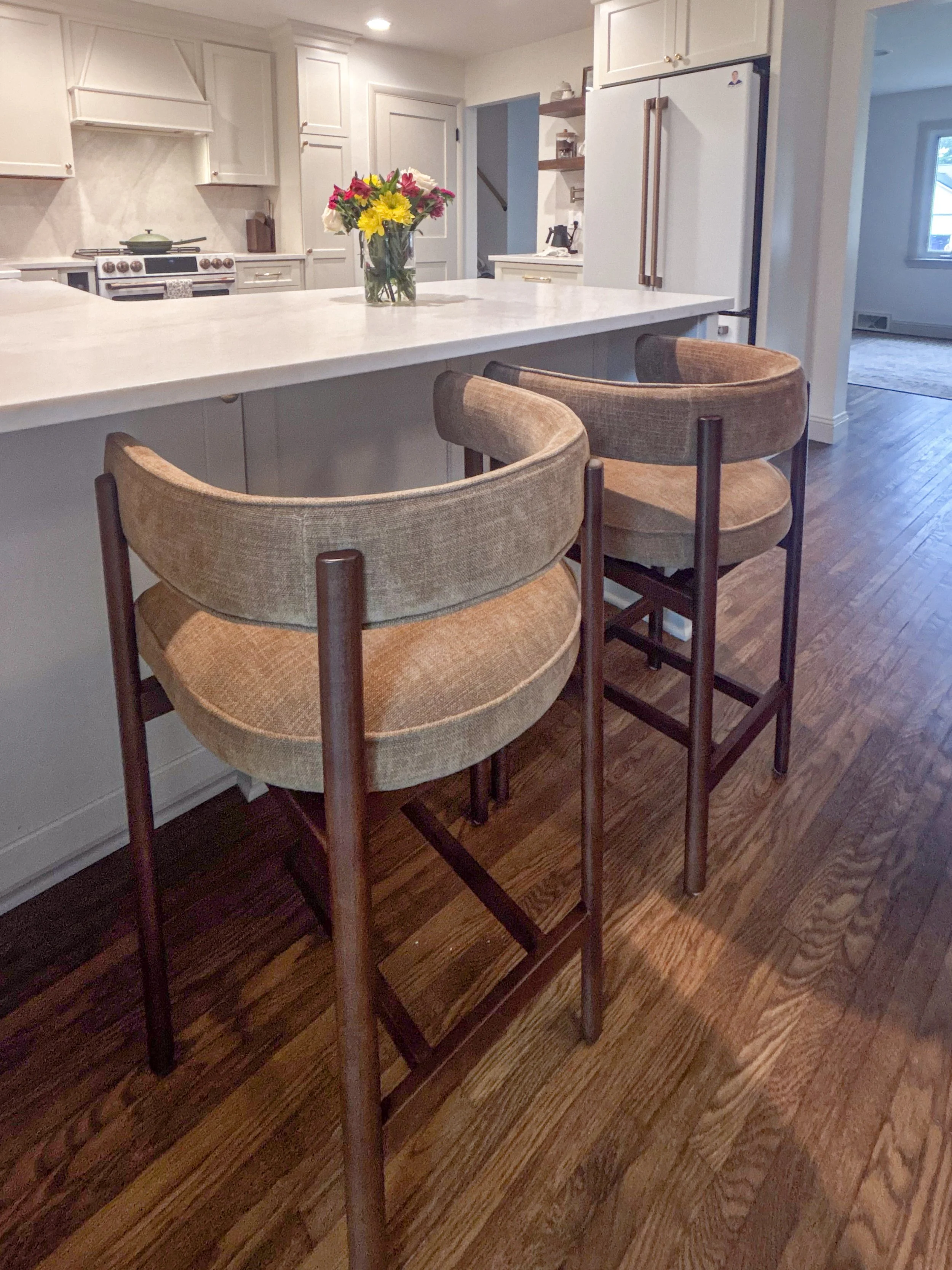 A kitchen interior with a white island countertop, beige upholstered bar stools, a vase with colorful flowers, white cabinetry, a stove with a green pot, a refrigerator, and hardwood flooring.