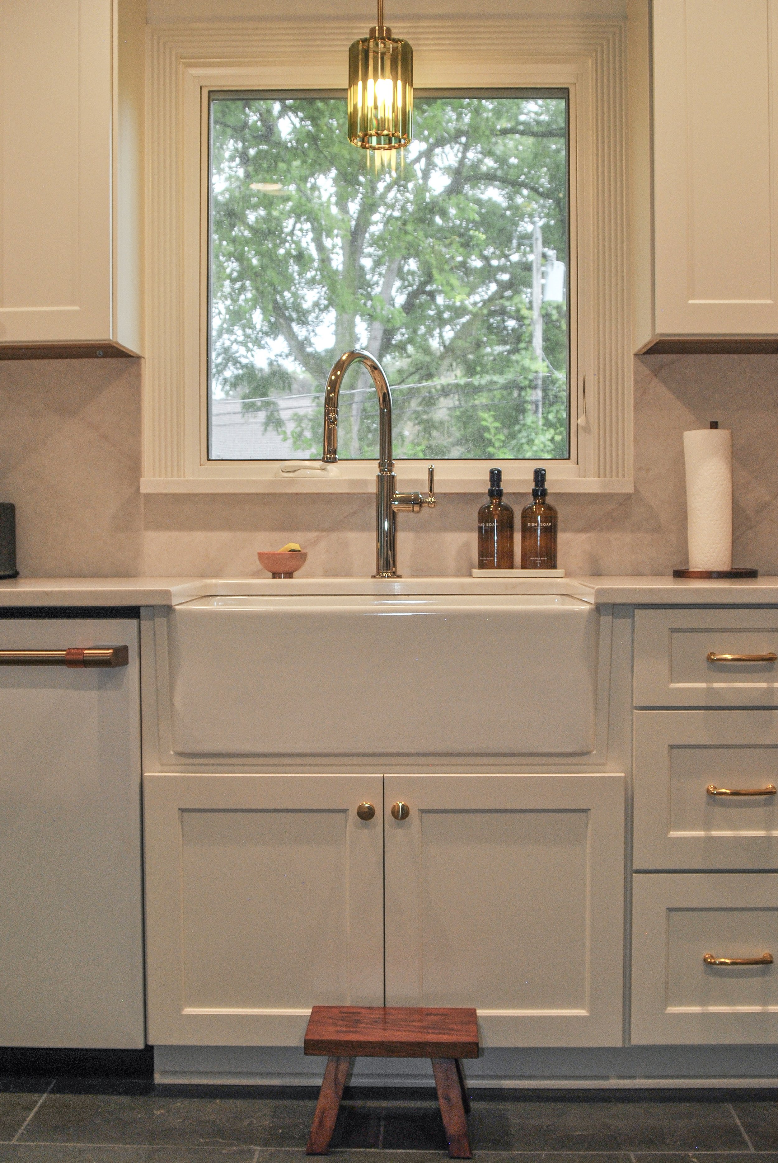 A kitchen with white cabinets, a countertop, a window with a green tree outside, a gold pendant light, a kitchen sink with a bronze faucet, two amber soap bottles, a paper towel roll, and a small wooden step stool on a dark tile floor.