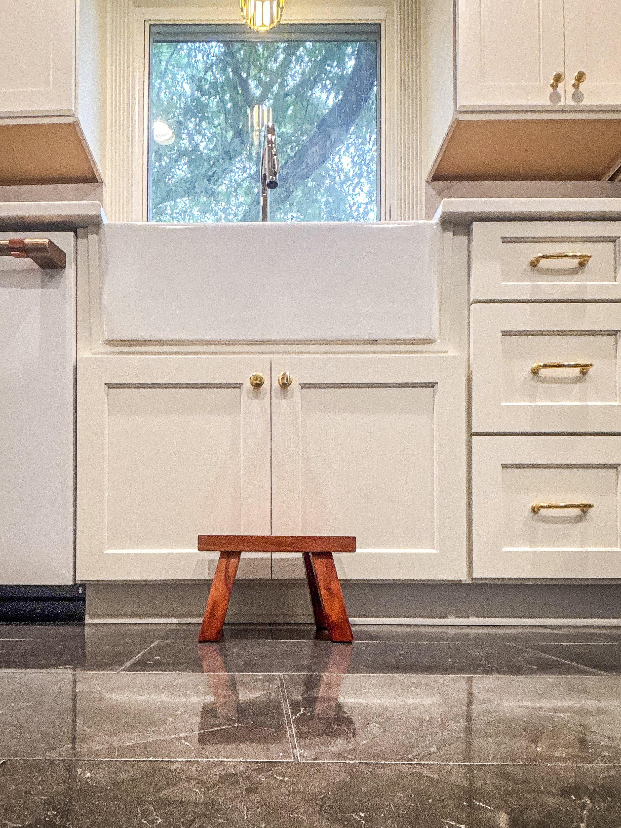 A kitchen with white cabinets, a farmhouse sink, a window with a view of trees outside, and a small wooden stool in front of the sink.