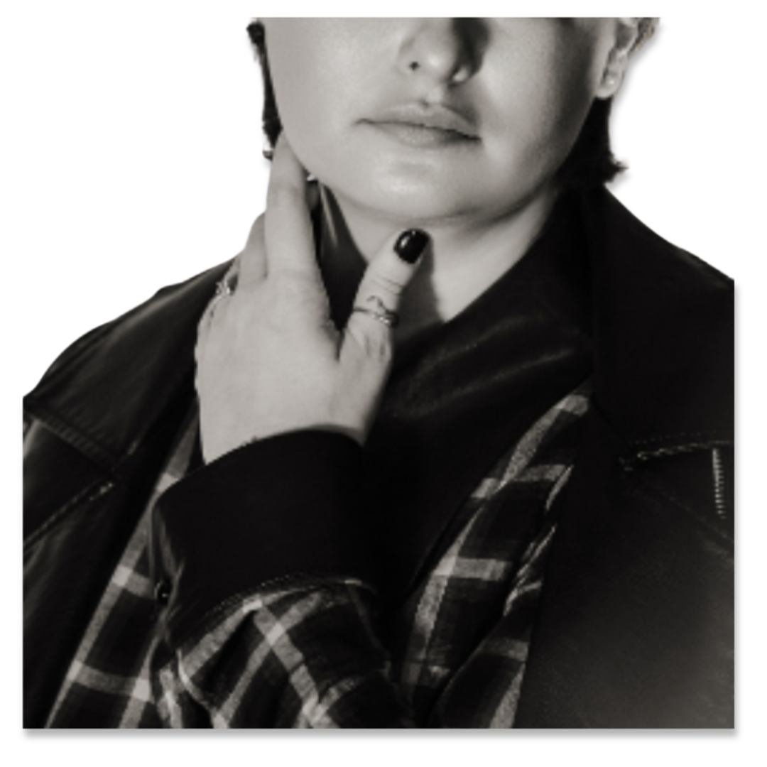 Close-up of a woman with short hair, wearing a leather jacket, plaid shirt, and rings, posing with her hand gently touching her neck against a black background.