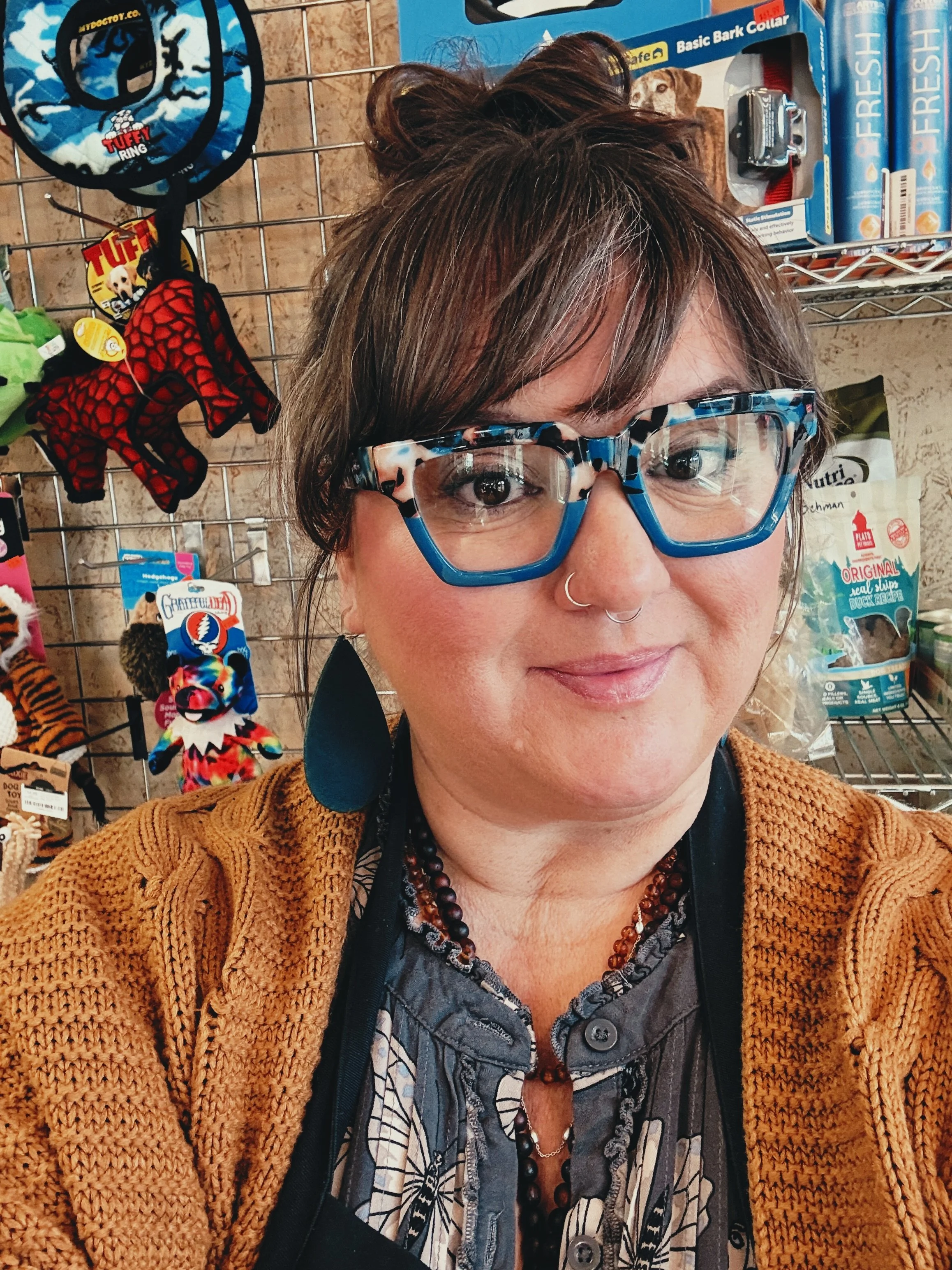 A woman with brown hair and glasses wearing a mustard yellow cardigan and a patterned shirt, standing in front of a store display of pet toys and supplies.