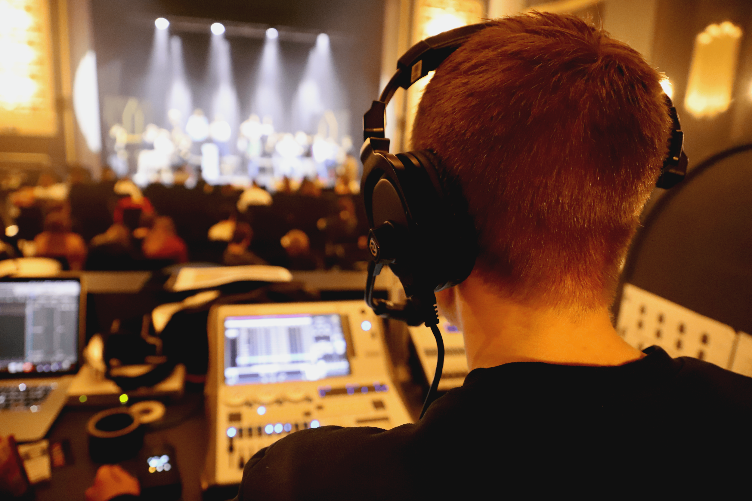 Lighting operator at the front-of-house position during a live performance at the Ritz Theatre, Penzance, monitoring the lighting desk with the stage and audience in the background.
