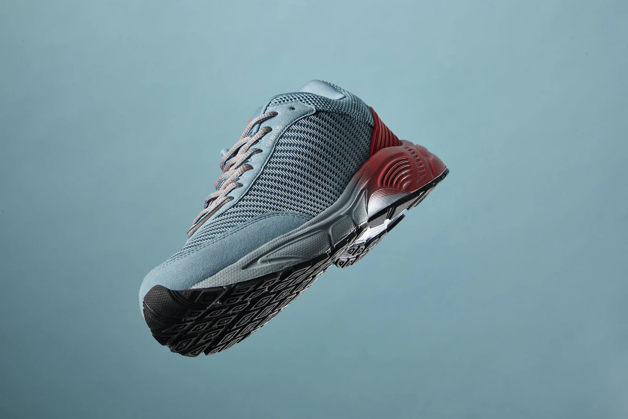A gray and red athletic running shoe suspended in mid-air against a light blue background.