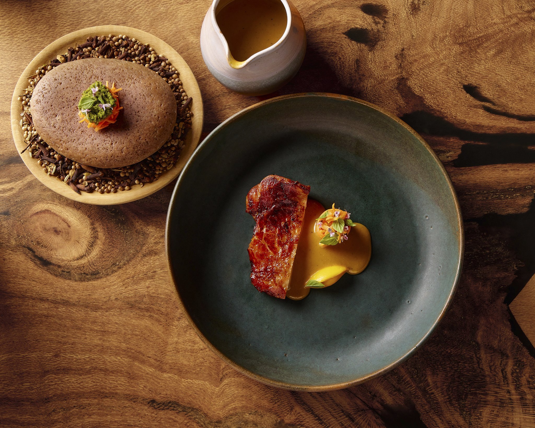 A wooden table with a ceramic pitcher, a dark green plate with a slice of glazed meat, and a wooden plate with a round bread roll garnished with green herbs and small edible flowers.