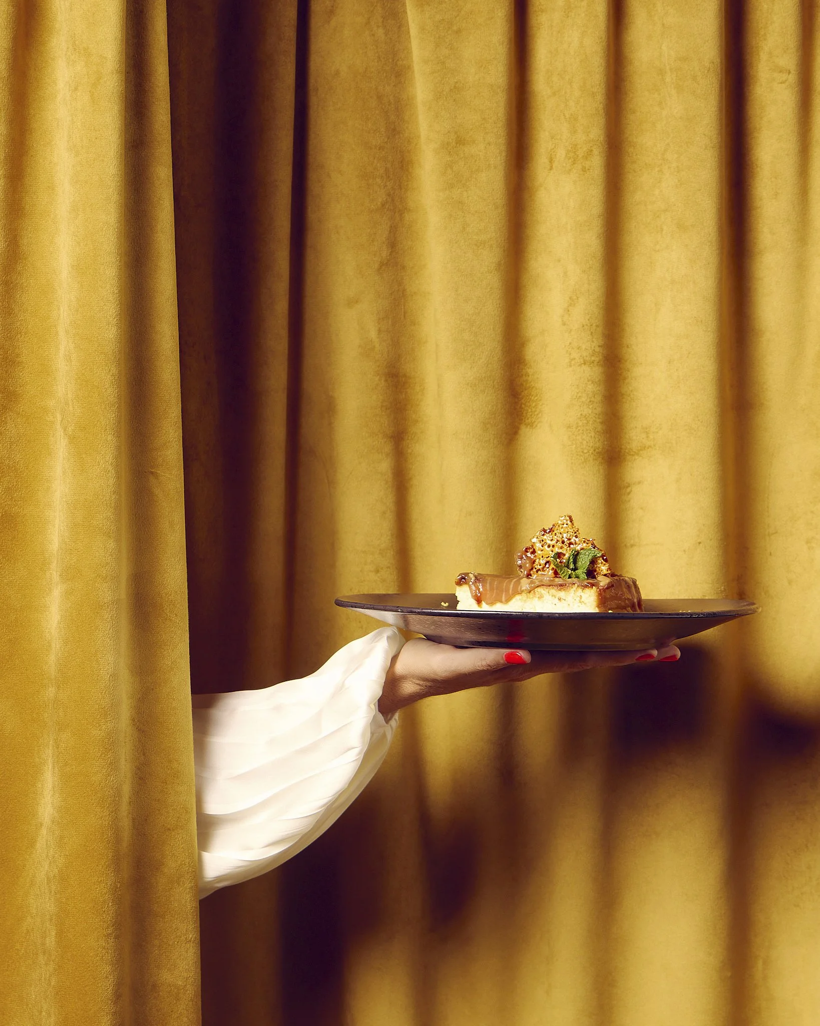 Person holding a black plate with a slice of cake topped with a decorated chocolate garnish and green mint leaves, against a gold curtain background.