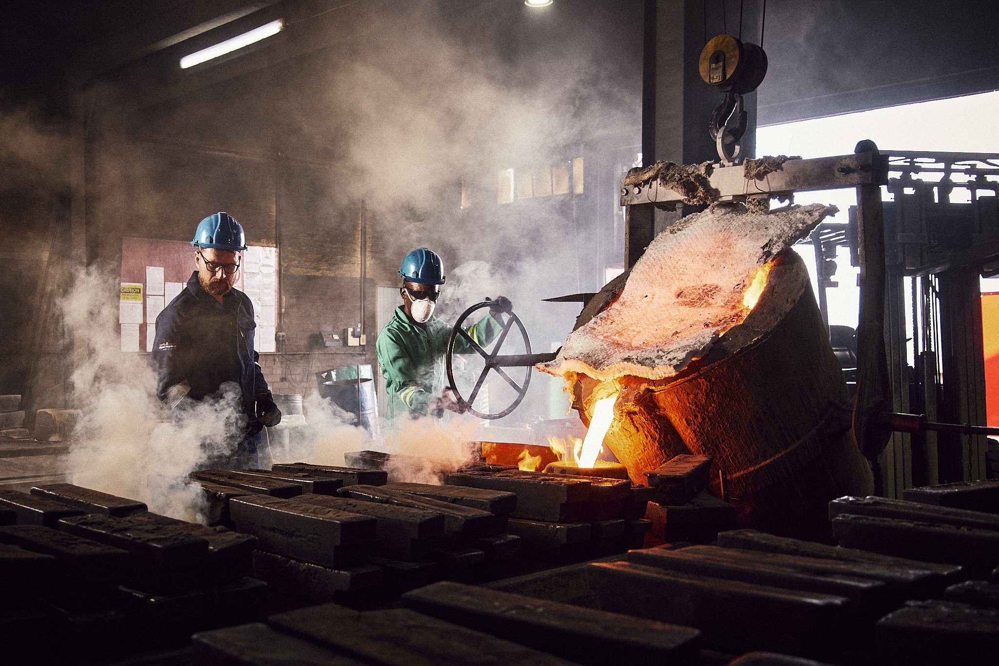 Two workers wearing safety helmets and masks are working with molten metal in an industrial foundry, with a large furnace pouring out bright orange-hot metal.