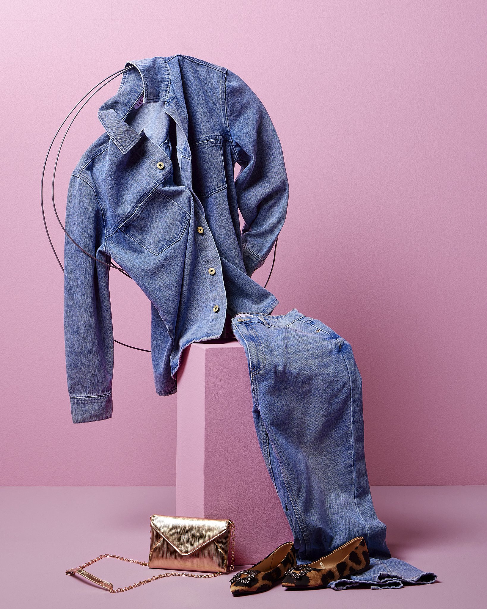 A denim jacket hanging on a black wire stand against a pink background. A matching pair of jeans and a pair of leopard print shoes with embellishments are below. A metallic rose gold clutch and chain strap purse are on the pink floor.