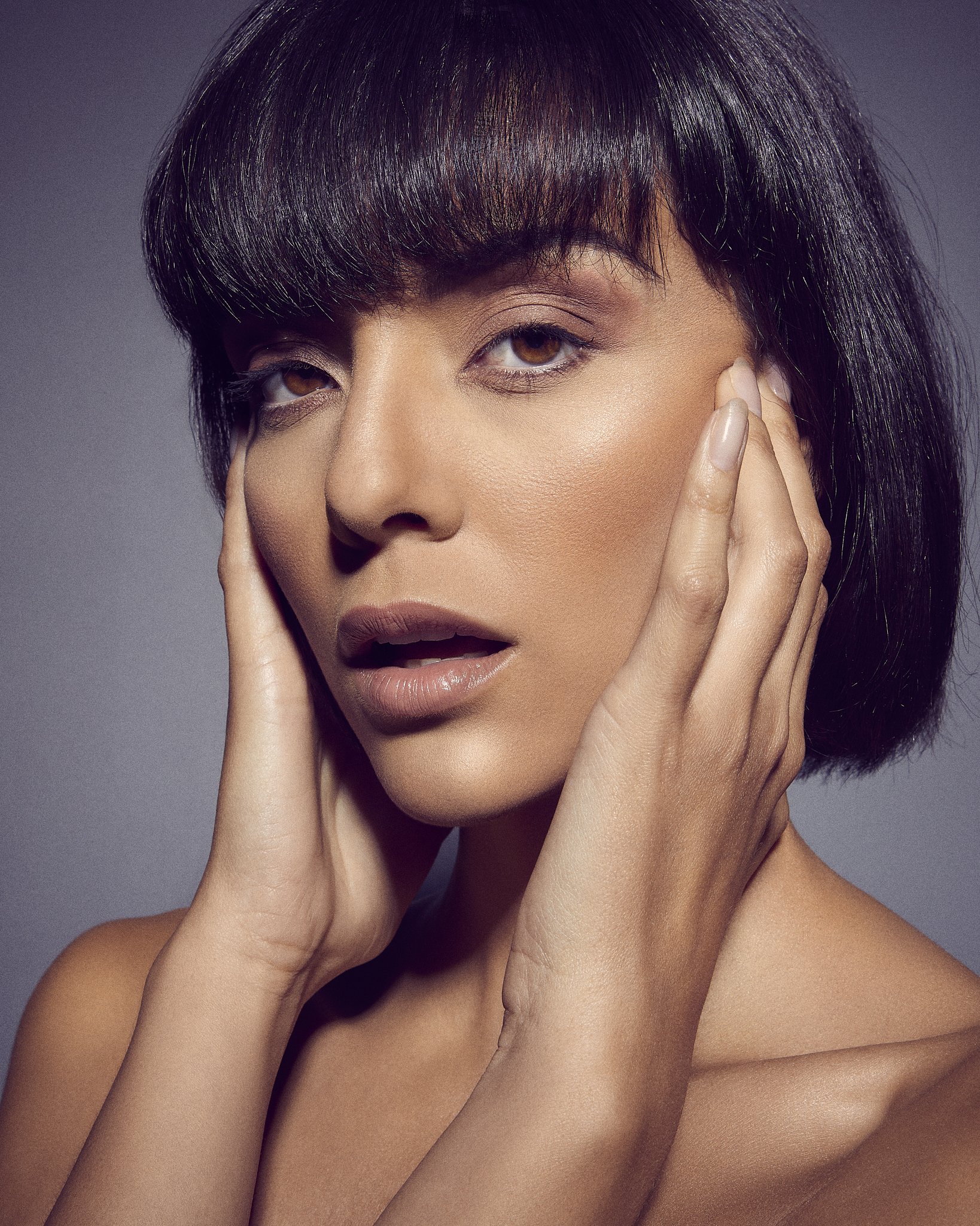 Close-up of a woman with dark brown hair and brown eyes touching her face with both hands, looking towards the camera.