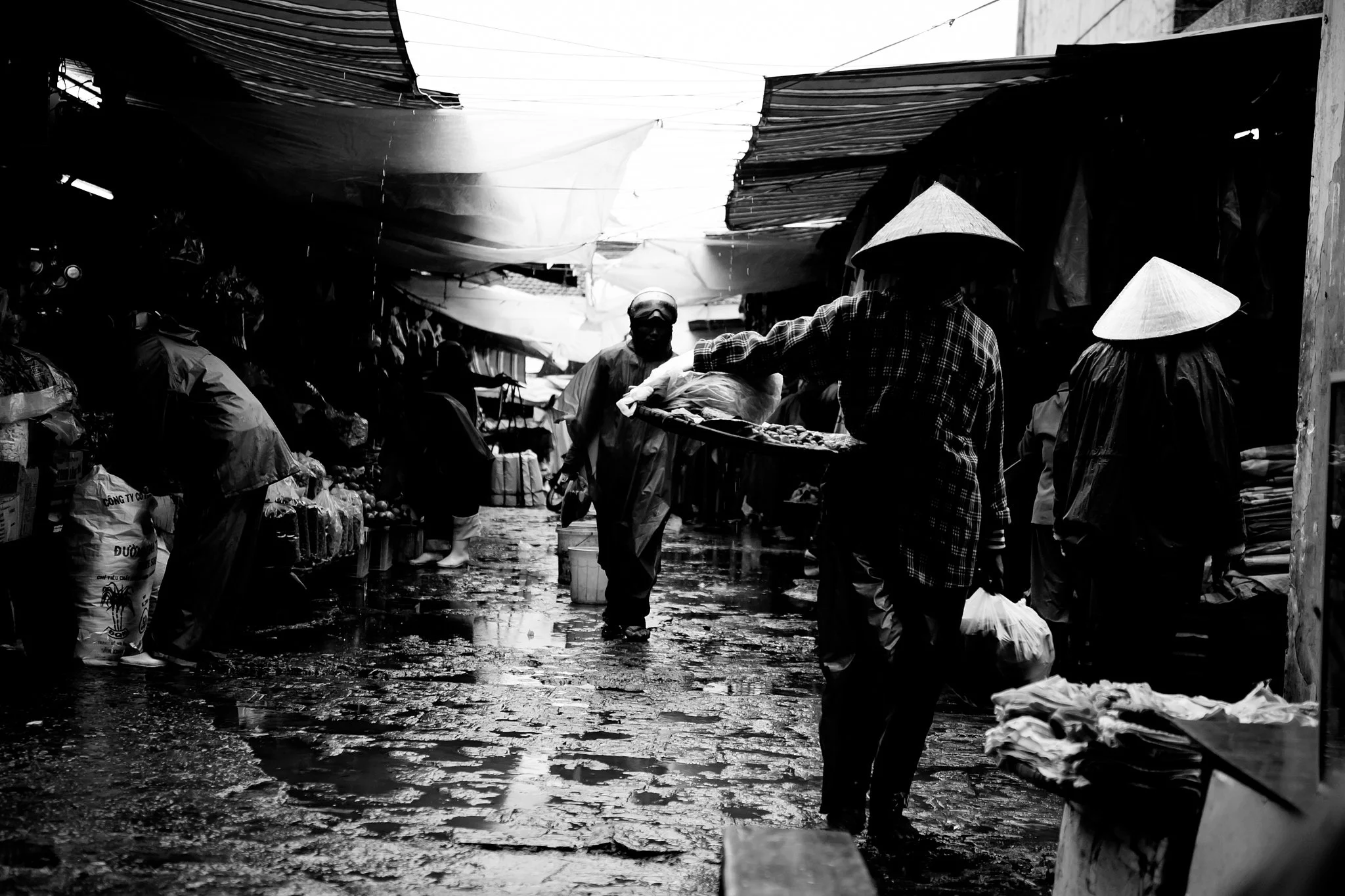 People shopping at the outdoor market in Hue, Vietnam with makeshift stalls, some individuals wearing conical hats, and the ground appears wet and muddy, possibly after rain.