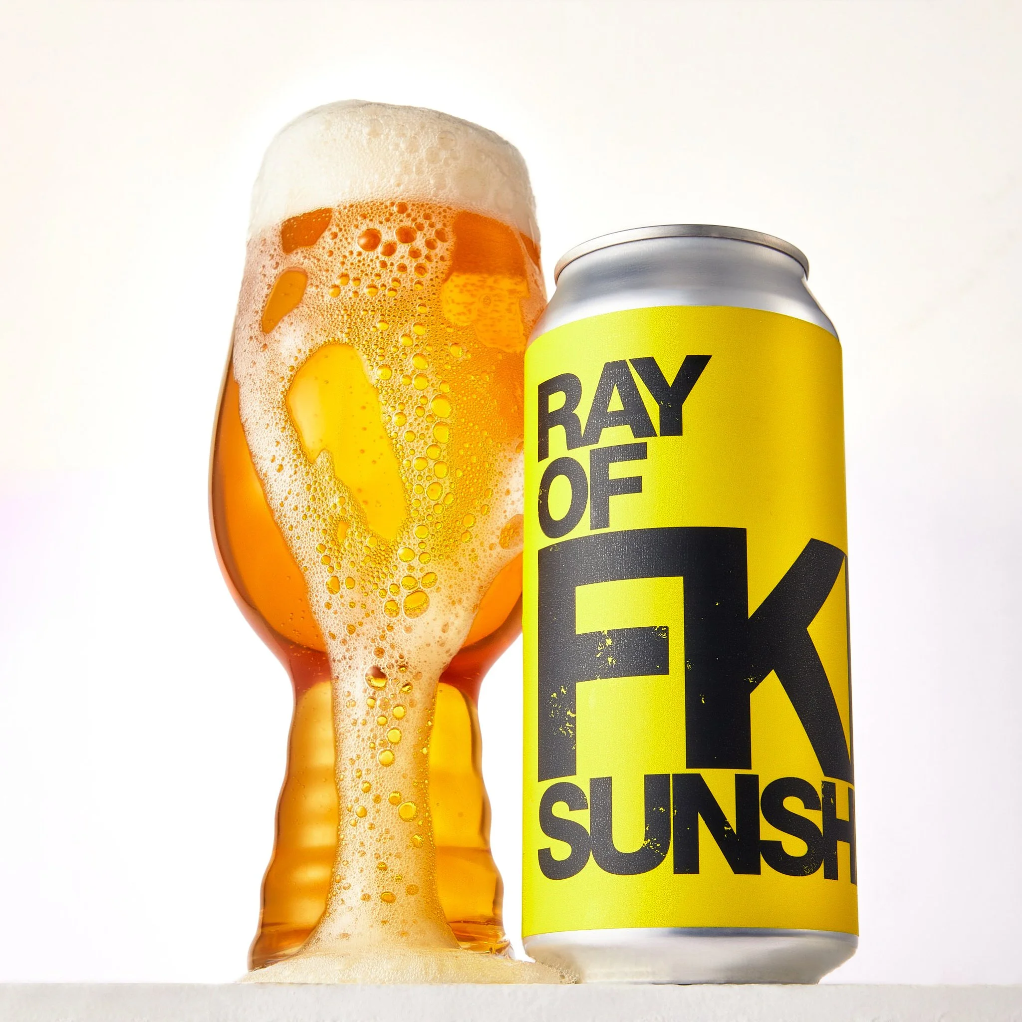 Close-up of a can of Ray of FKN Sunshine, with a yellow label, and a glass of orange-colored freshly poured beer with a foamy head, placed on a white surface against a white background.