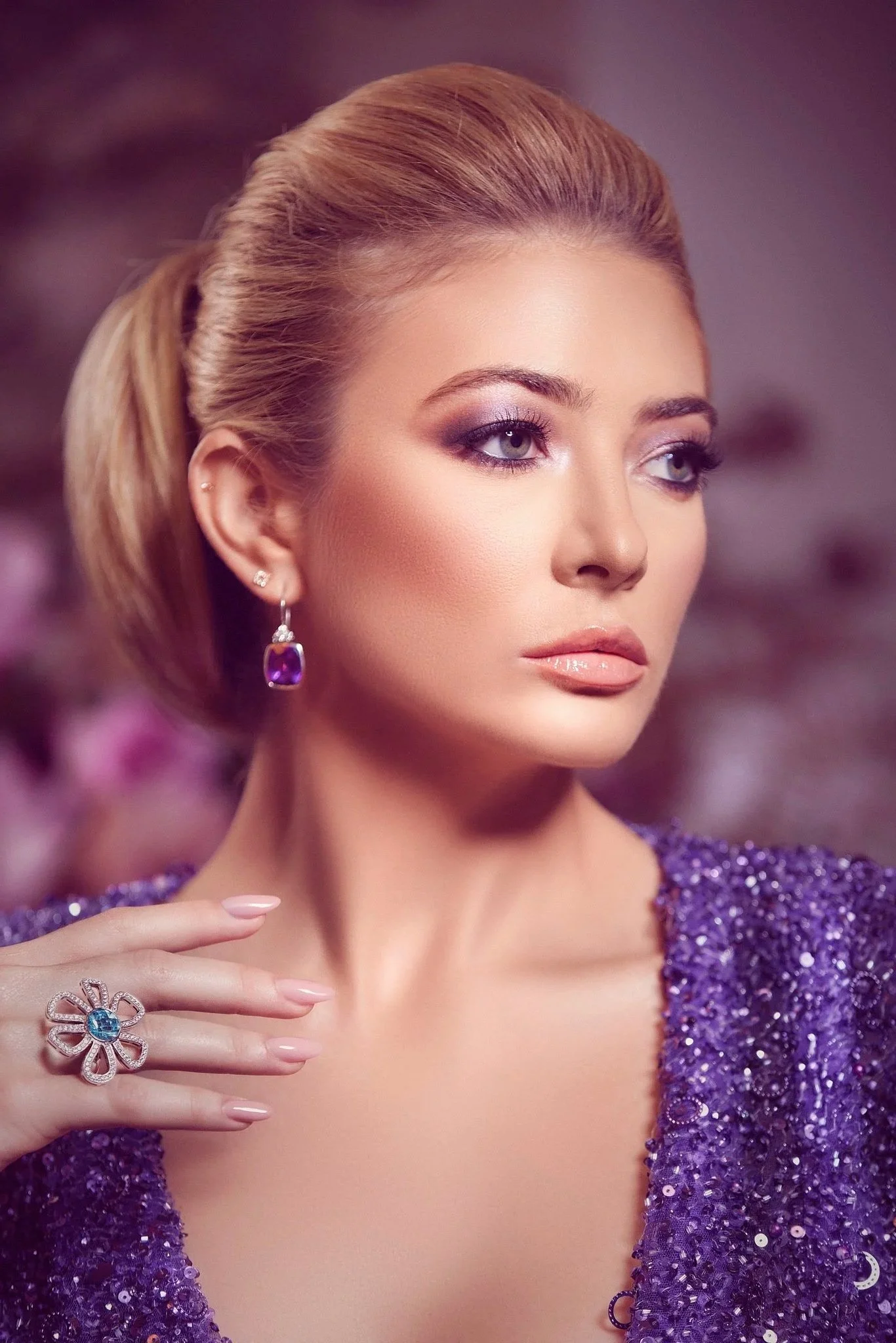 Close-up of a woman with blonde hair in an elegant updo, wearing purple earrings and a purple sparkly dress, with makeup emphasizing her eyes and lips, and a large flower-shaped ring with a blue gemstone on her finger.