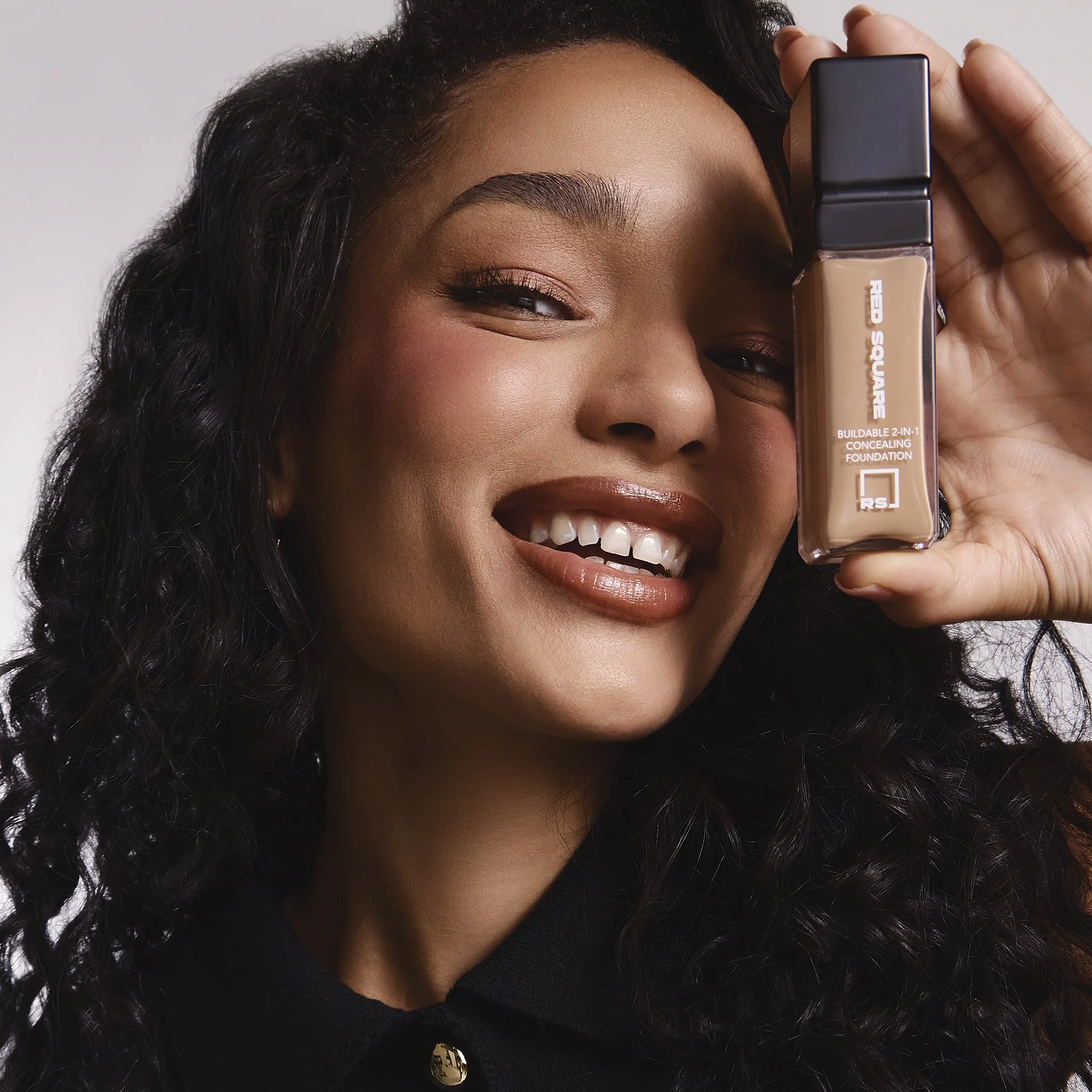 A woman with dark, curly hair smiling while holding a bottle of foundation close to her face.