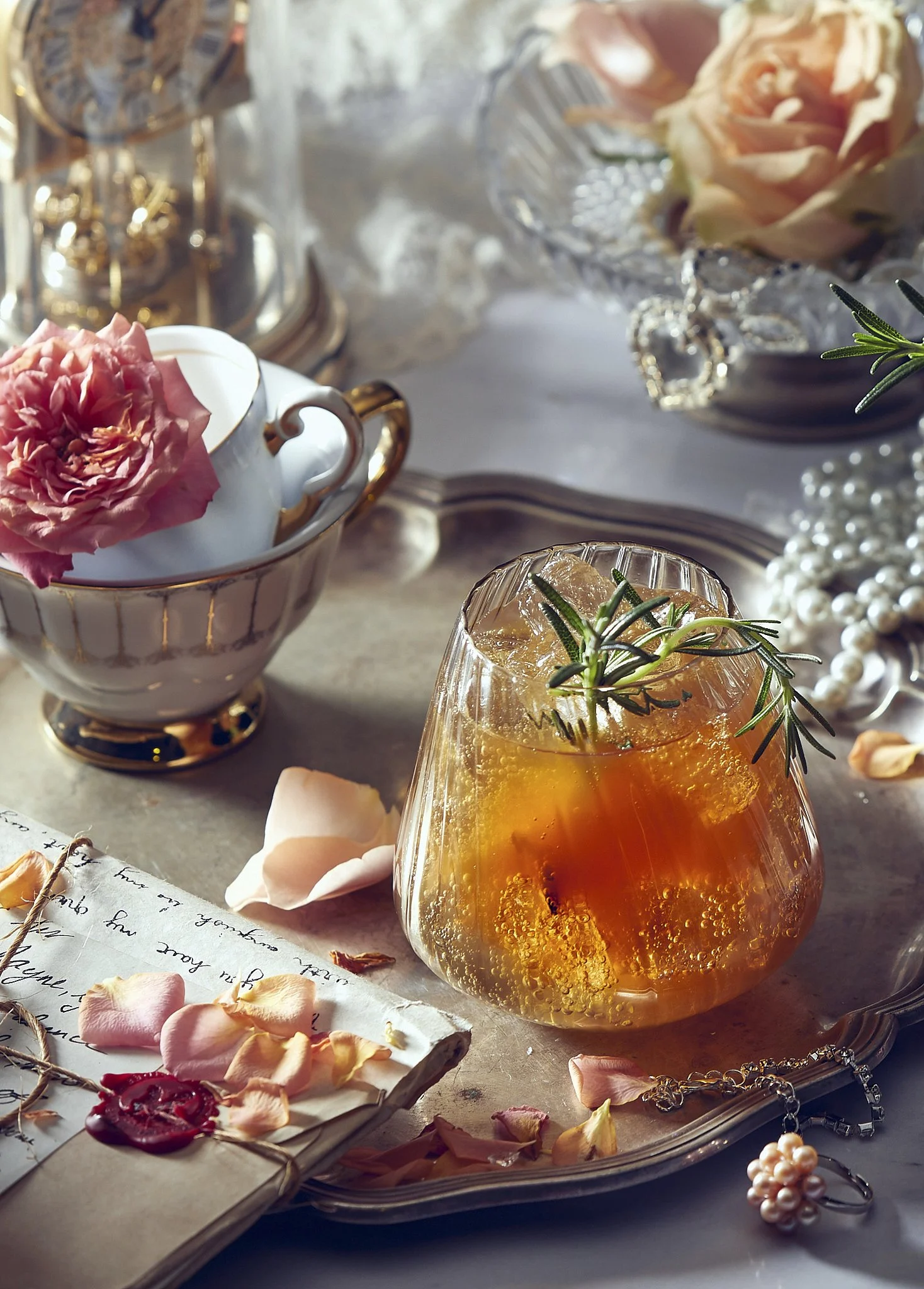 A close-up of a glass of iced tea with sprigs of rosemary, surrounded by pink rose petals, jewelry, and decorative items on a silver tray.