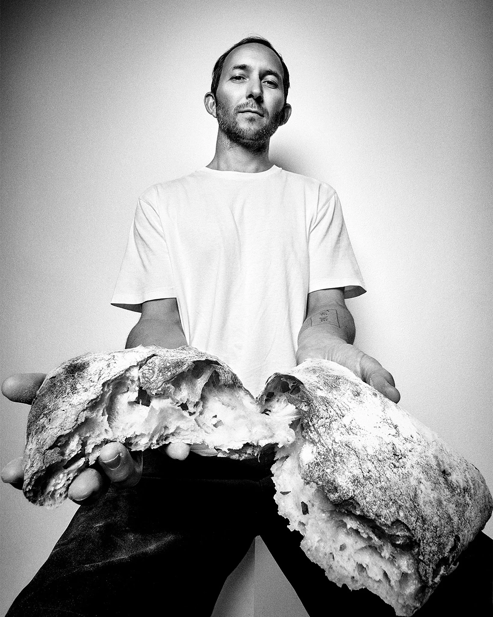 super wide angle Black and white photo of a man holding a large, torn open loaf of bread, standing in front of a plain backdrop.