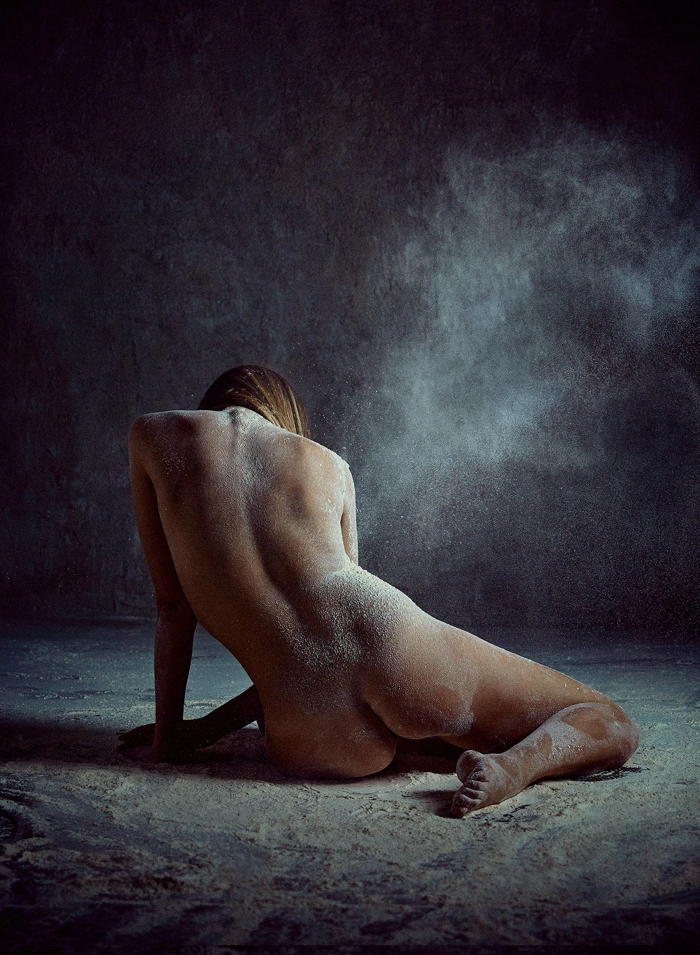 A woman sitting on the ground with her back to the camera, covered in white powder or chalk, in a dark setting with a mist or fog in the background.