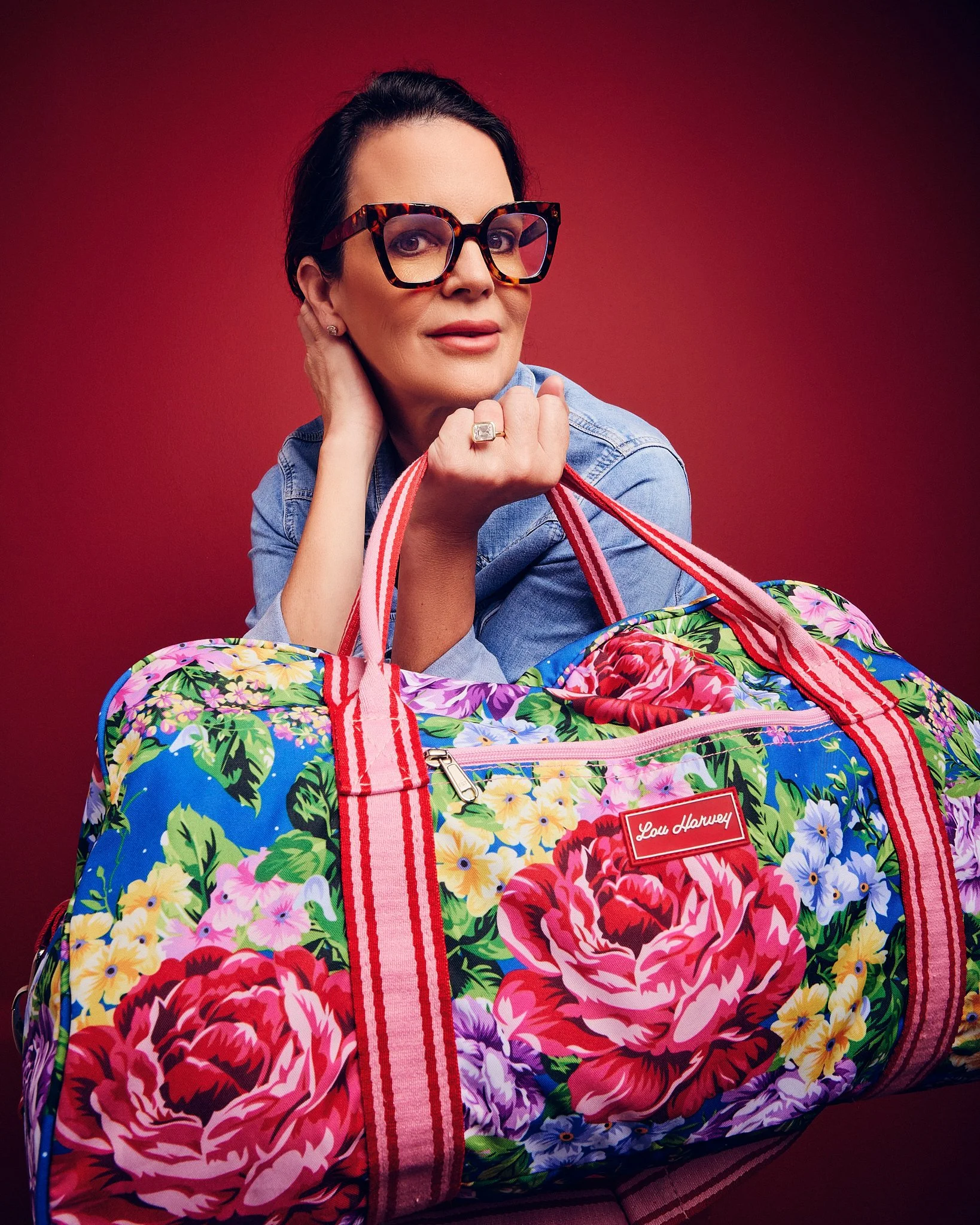 A woman with dark hair, wearing large tortoiseshell glasses, a silver earring, and a denim jacket, is resting her head on her hand. She is sitting behind a colorful floral duffle bag with striped straps and a red label that reads 'Lou Harvey.' The ba