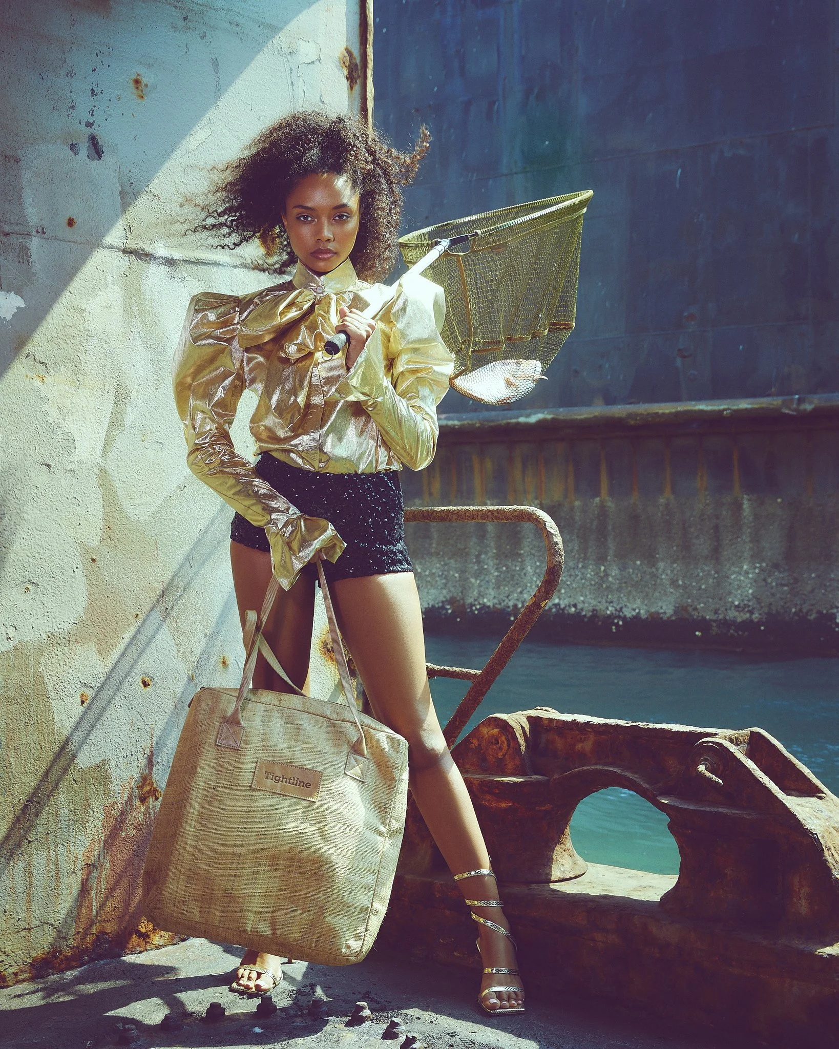 A woman with curly hair stands against a rustic wall at the seashore, holding a floating net and wearing a shiny gold jacket, a black sequined skirt, and silver high-heeled sandals, with a beige tote bag labeled 'Tightline' beside her.