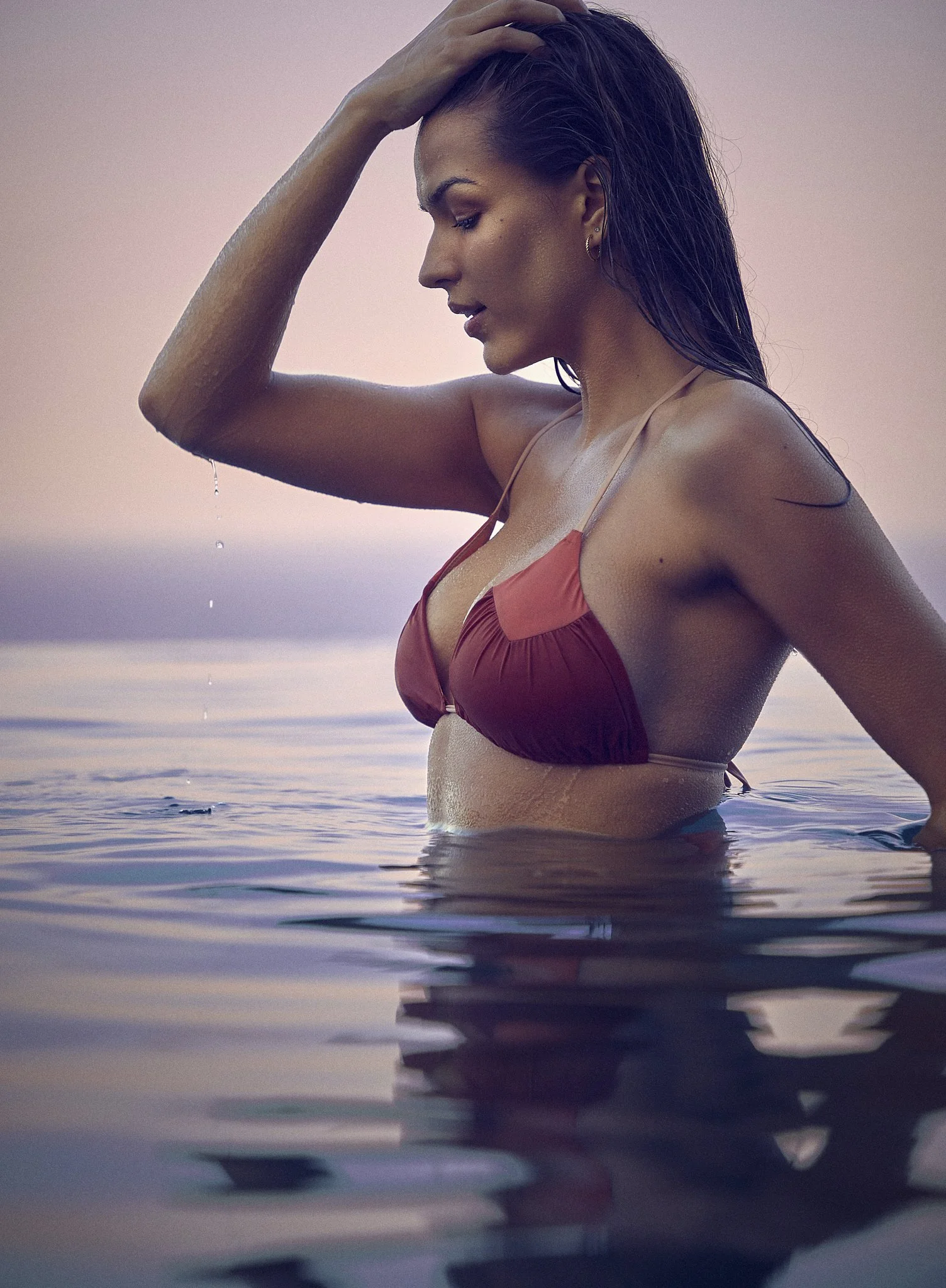 Woman in a red bikini standing in water, touching her wet hair, during sunset or sunrise with calm water and soft sky in the background.