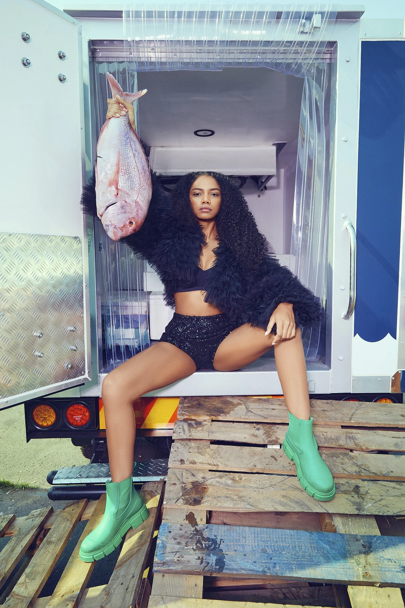 A woman with curly hair seated on a wooden pallet platform in the back of a truck, holding a large pink fish, wearing a black outfit with ruffled sleeves and bright green rubber boots.