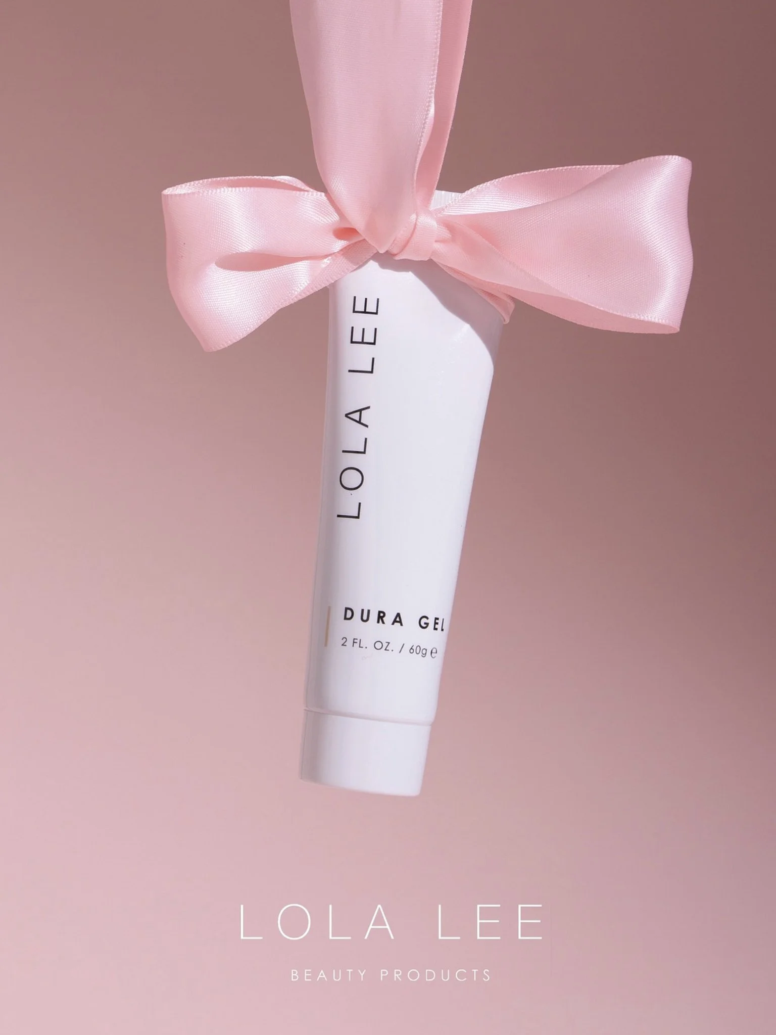 A white tube of Lola Lee Dura Gel with a pink satin ribbon tied around it, set against a pink background, with text at the bottom reading 'Lola Lee Beauty Products'.