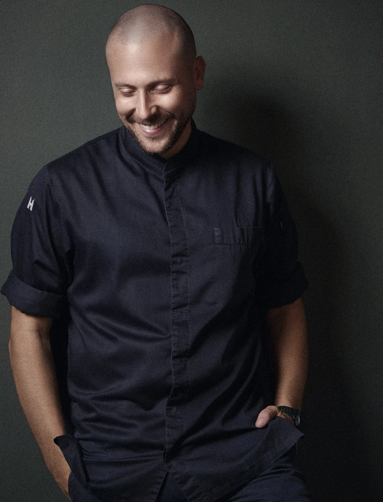 Johannes Richter, chef at The Livingroom  is smiling and looking down, wearing a black button-up shirt, sitting against a dark green background.