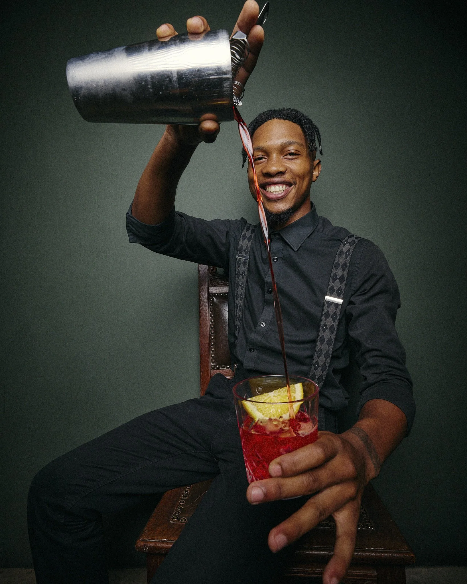 A smiling man in a black shirt and suspenders pours red liquid from a metal pitcher into a glass filled with ice, lemon slice, and a cherry, while sitting on a wooden chair against a dark green background.