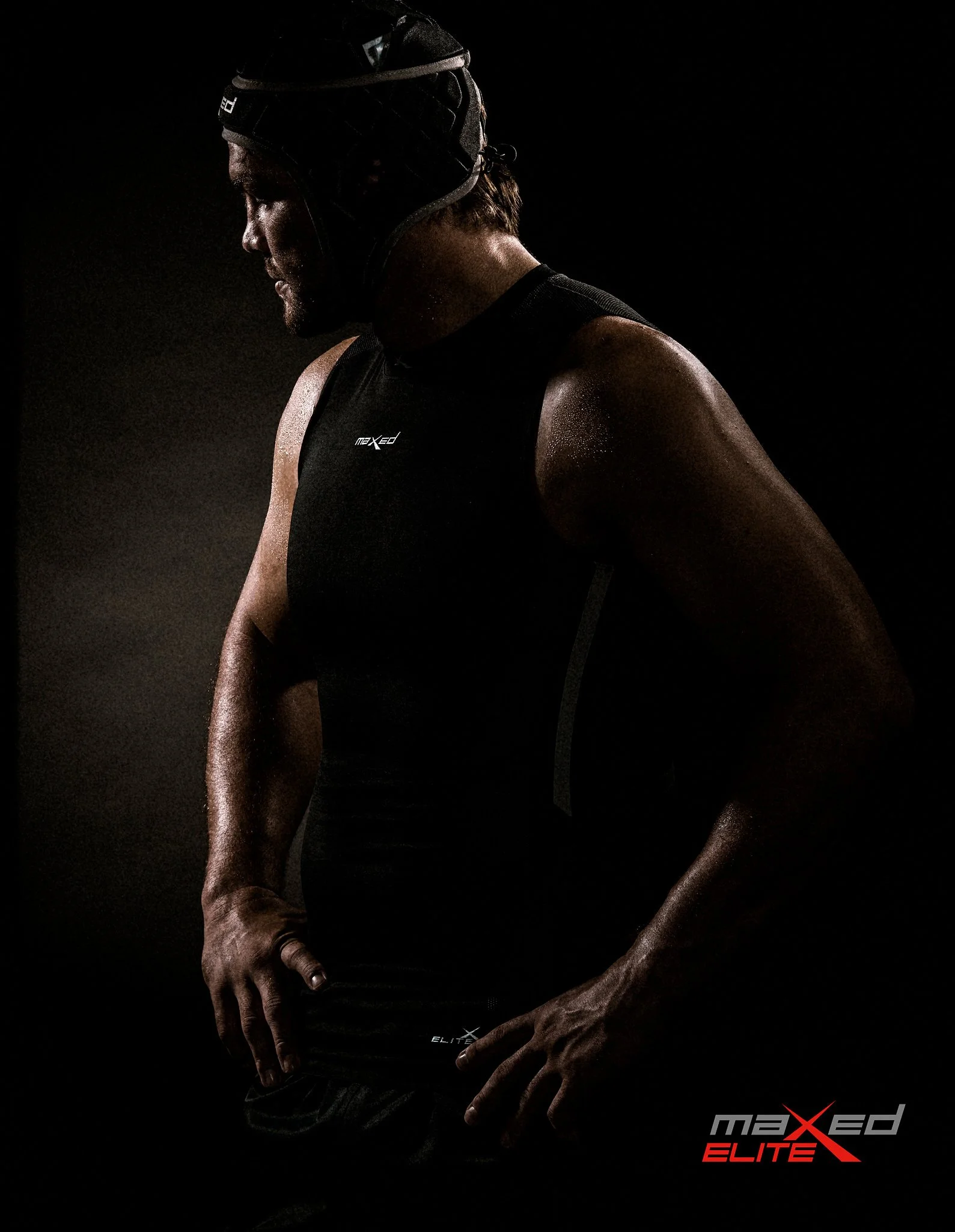 Flip van der Merwe wearing a black Maxed Eltie sports shirt and a black head covering, standing with his head bowed and his right hand resting on his lower back, illuminated against a dark background. Rugby legend