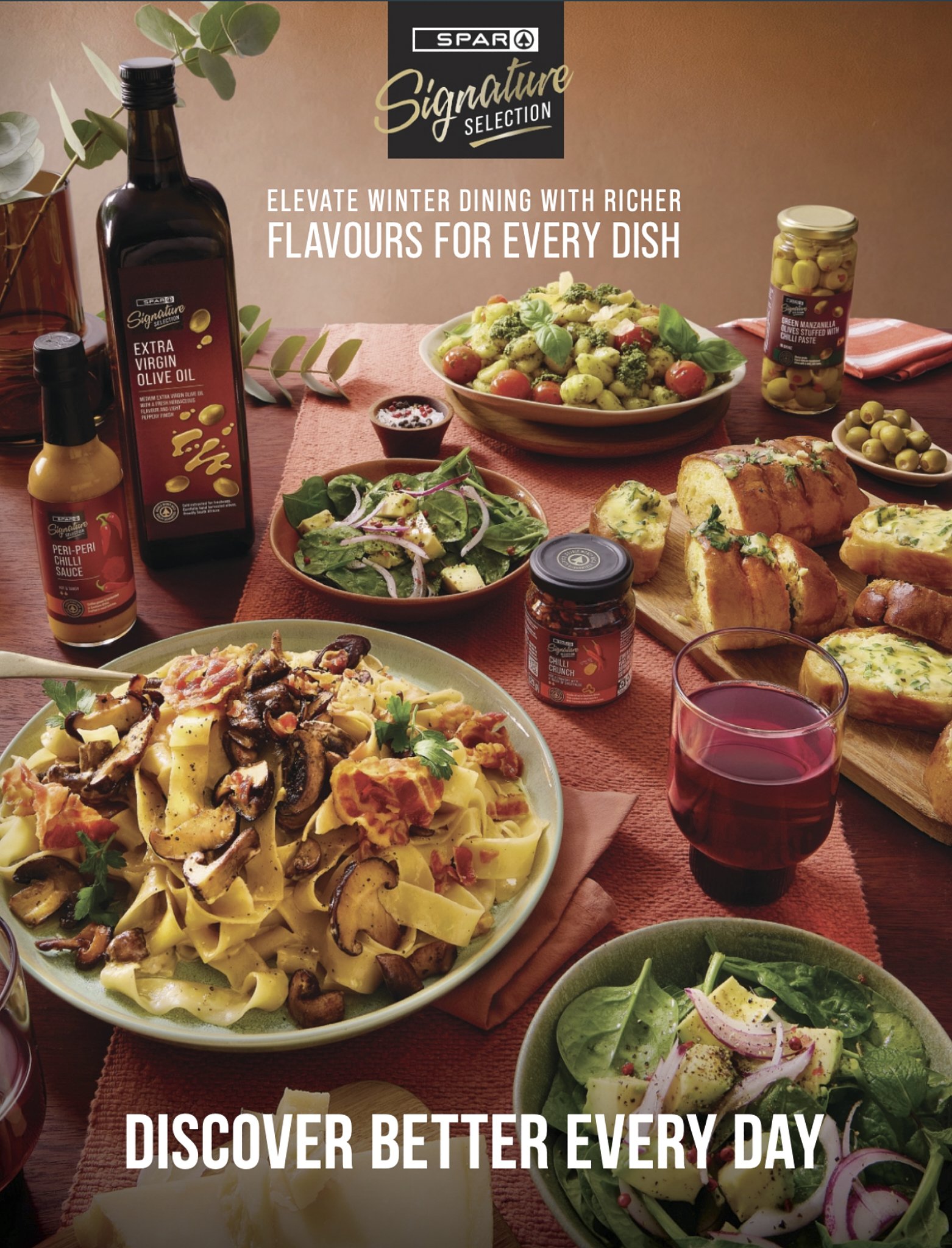 A table with various dishes including pasta with mushrooms and tomatoes, salad with spinach and onions, and a bowl of chopped vegetables, along with bottles of olive oil and chili sauce, and glasses of red wine, set on a table with a peach-colored ta