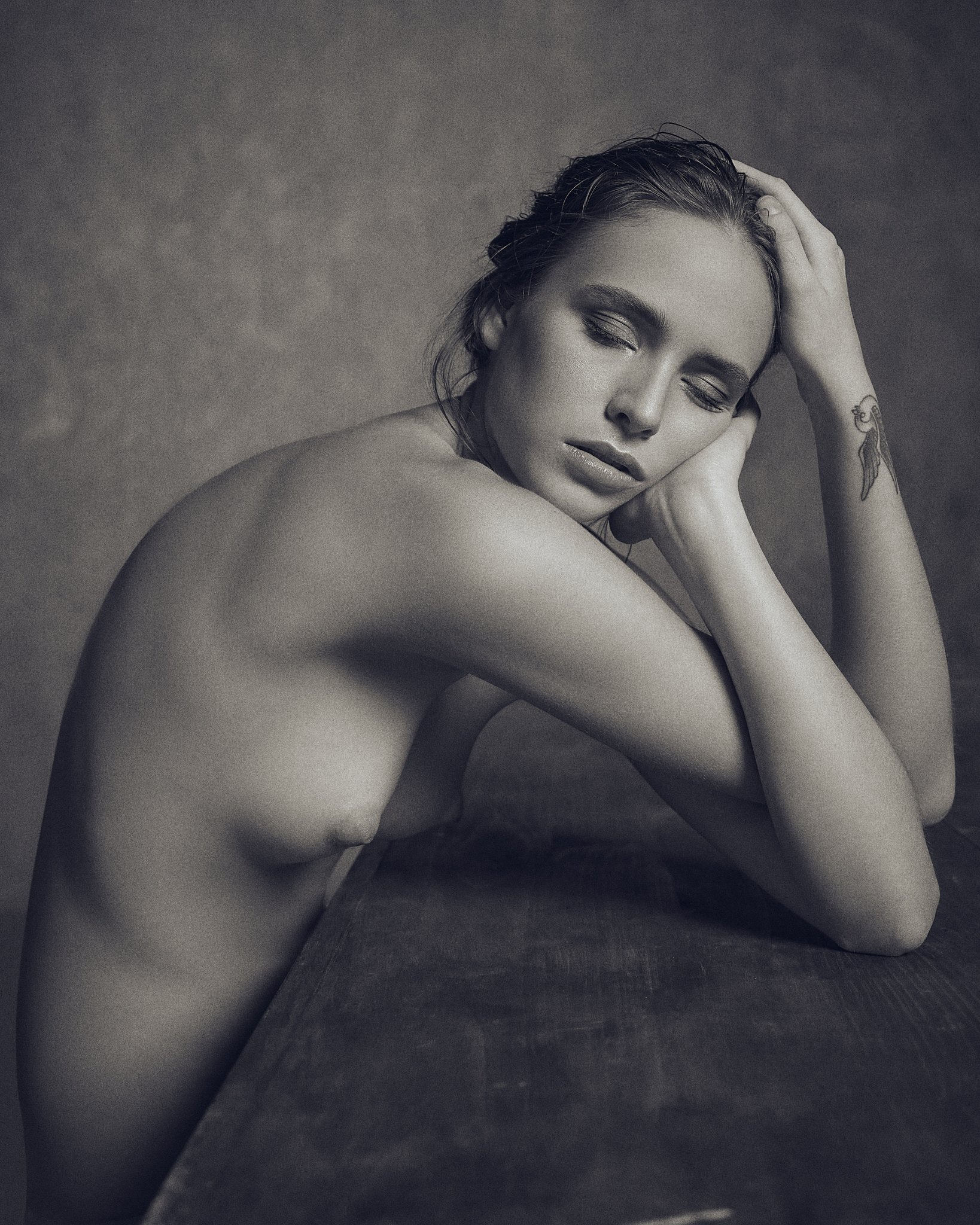 A nude woman with wet hair, eyes closed, resting her head on her hand with a tattoo of a swallow on her arm, posing against a gradient background.
