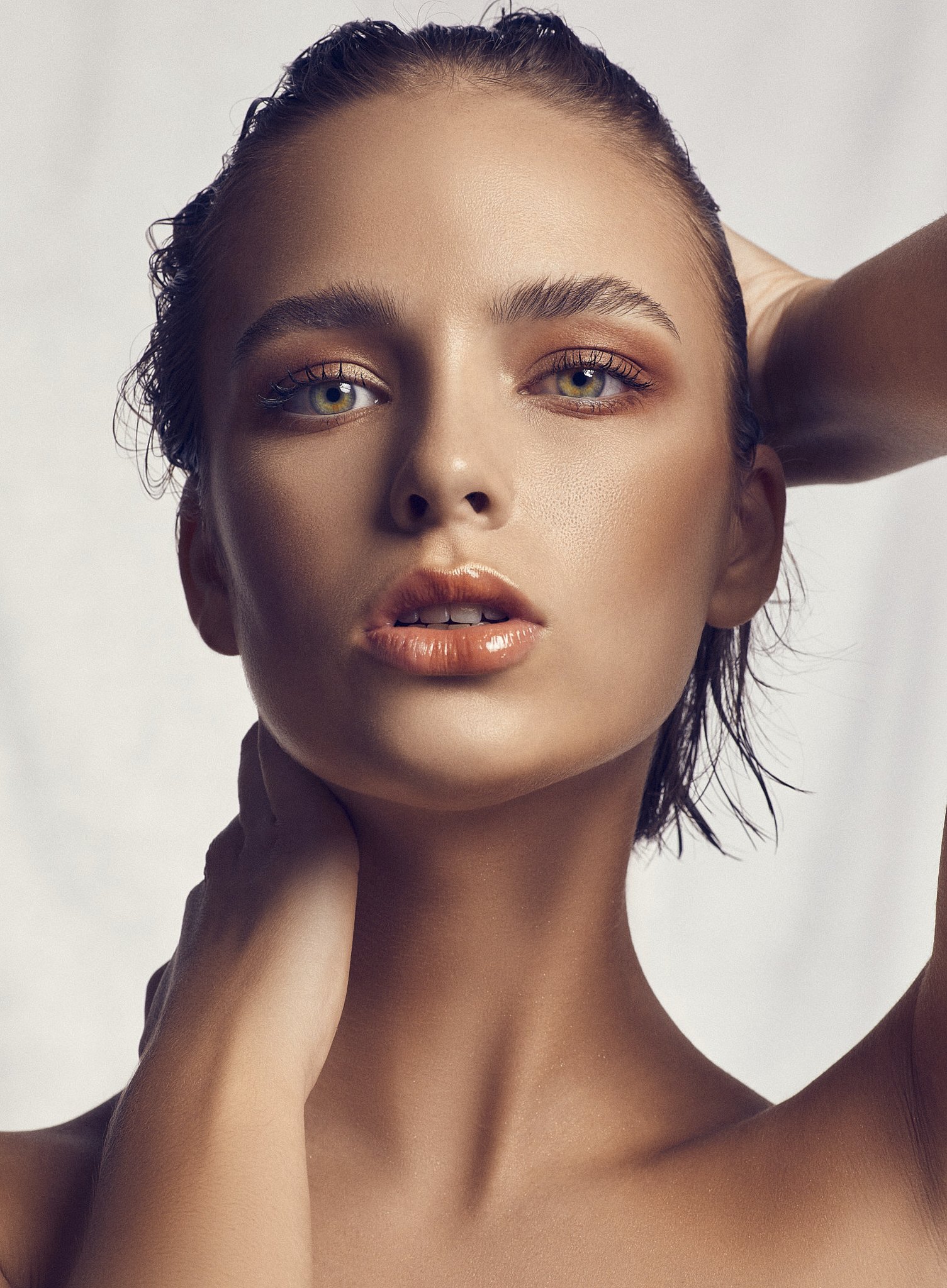 A close-up portrait of a woman with wet hair, light makeup, and piercing eyes, posing with her hand behind her head against a plain light background.