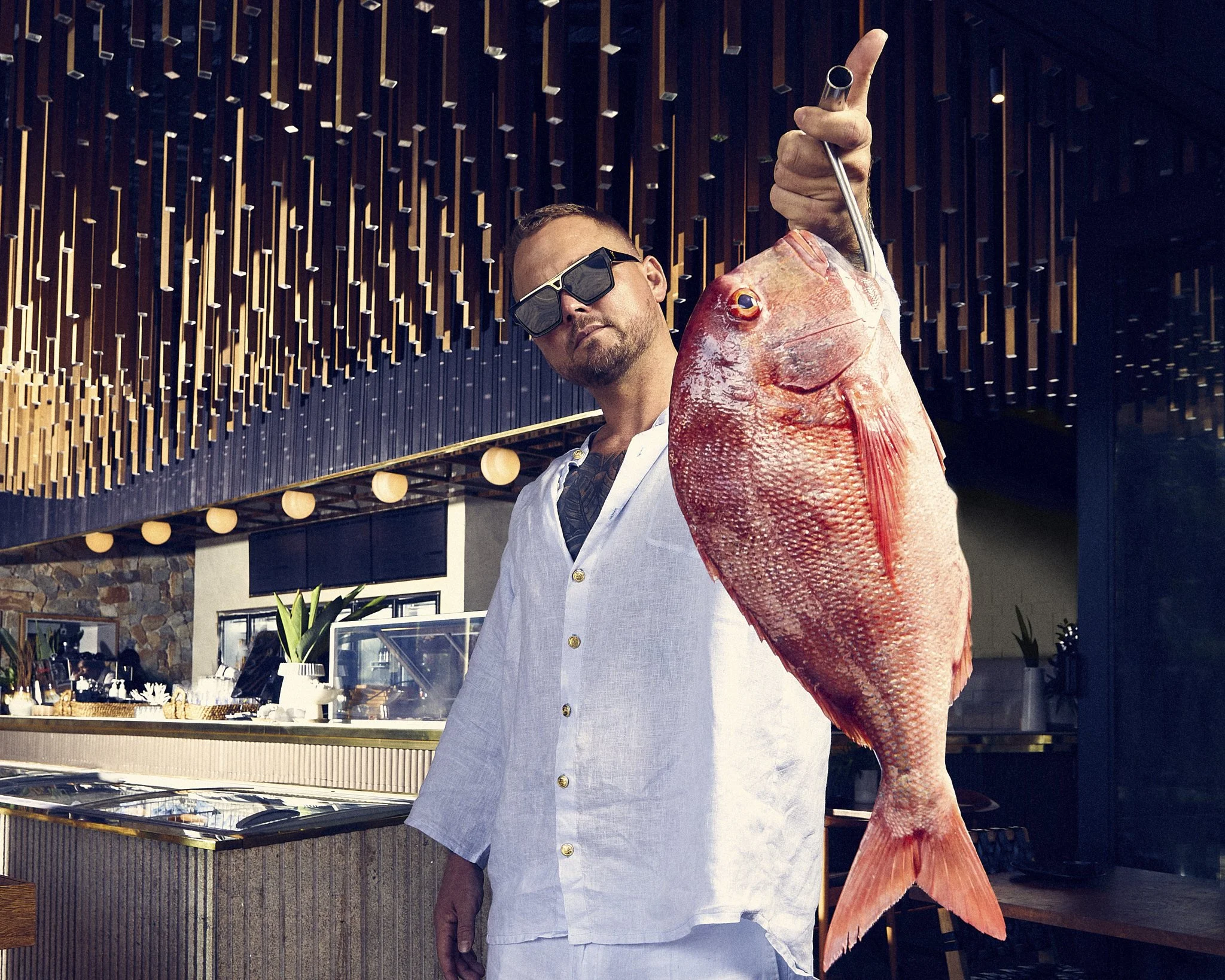 Tyler from Tightline wearing sunglasses and white shirt holding a large red fish in Tightline restaurant or cafe setting.