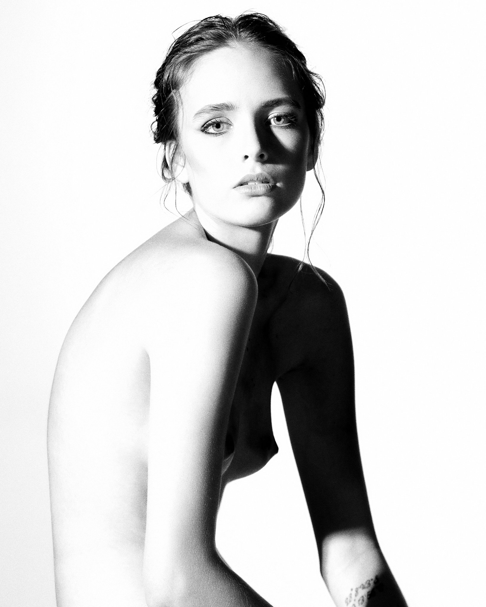 Black and white portrait of a woman with light-colored eyes, wet hair, and part of a tattoo visible on her arm, looking directly at the camera.