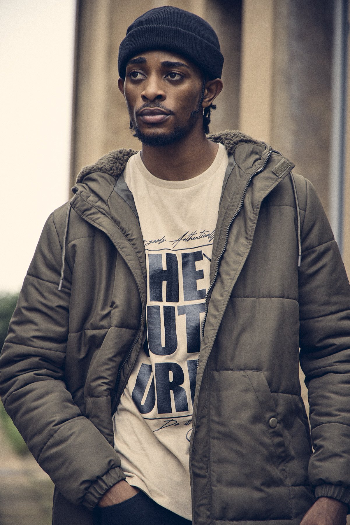 A young man wearing a black beanie, a beige graphic t-shirt, and a greenpuffer jacket stands outdoors.