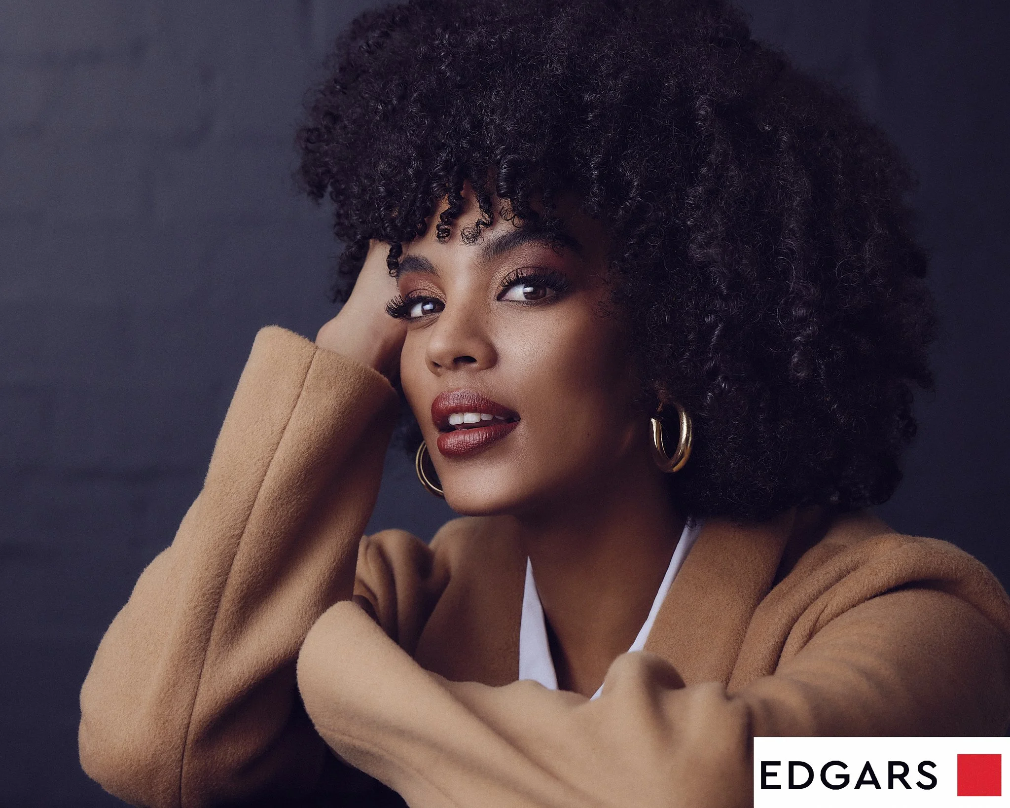 Close-up of a woman with curly hair, gold hoop earrings, wearing a tan coat and white top, posing against a dark background.