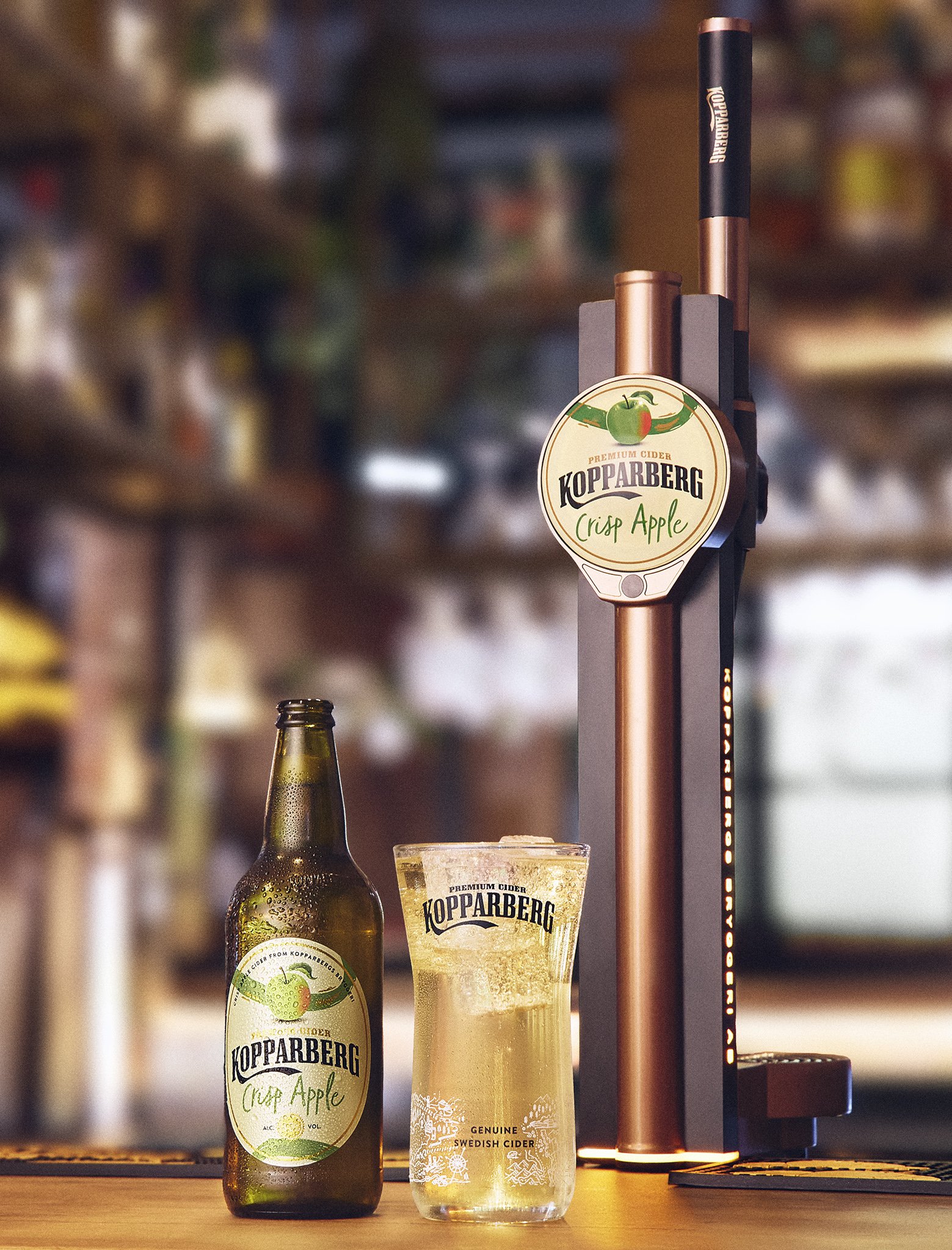 A bottle of Kopparberg Crisp Apple cider, a glass of cider, and a cask tap in a bar setting.