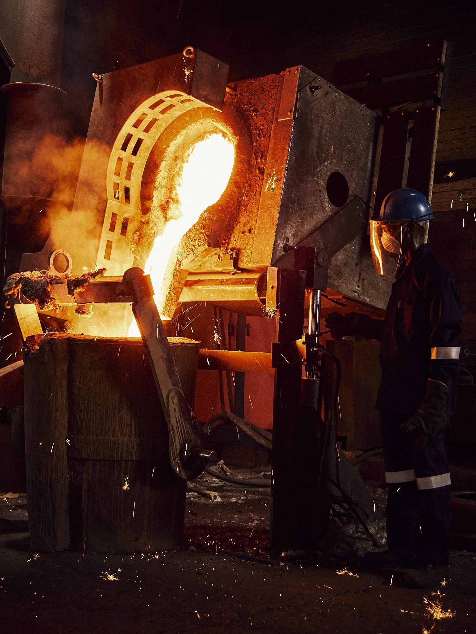 Blacksmith pouring molten metal from a large furnace with bright flames, sparks flying, and a worker observing while wearing protective gear.