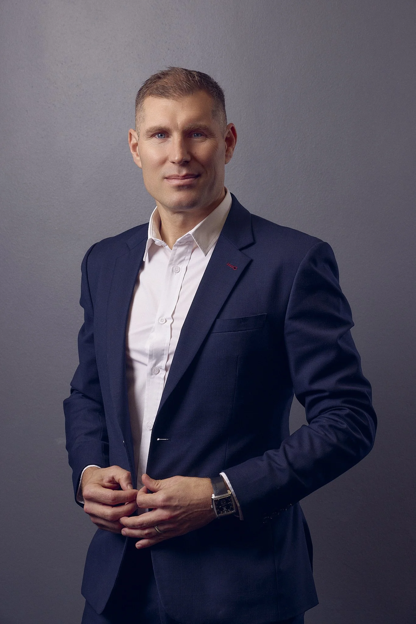 A man in a dark navy suit and white shirt stands against a plain gray background, looking at the camera, with his hands clasped in front of him.