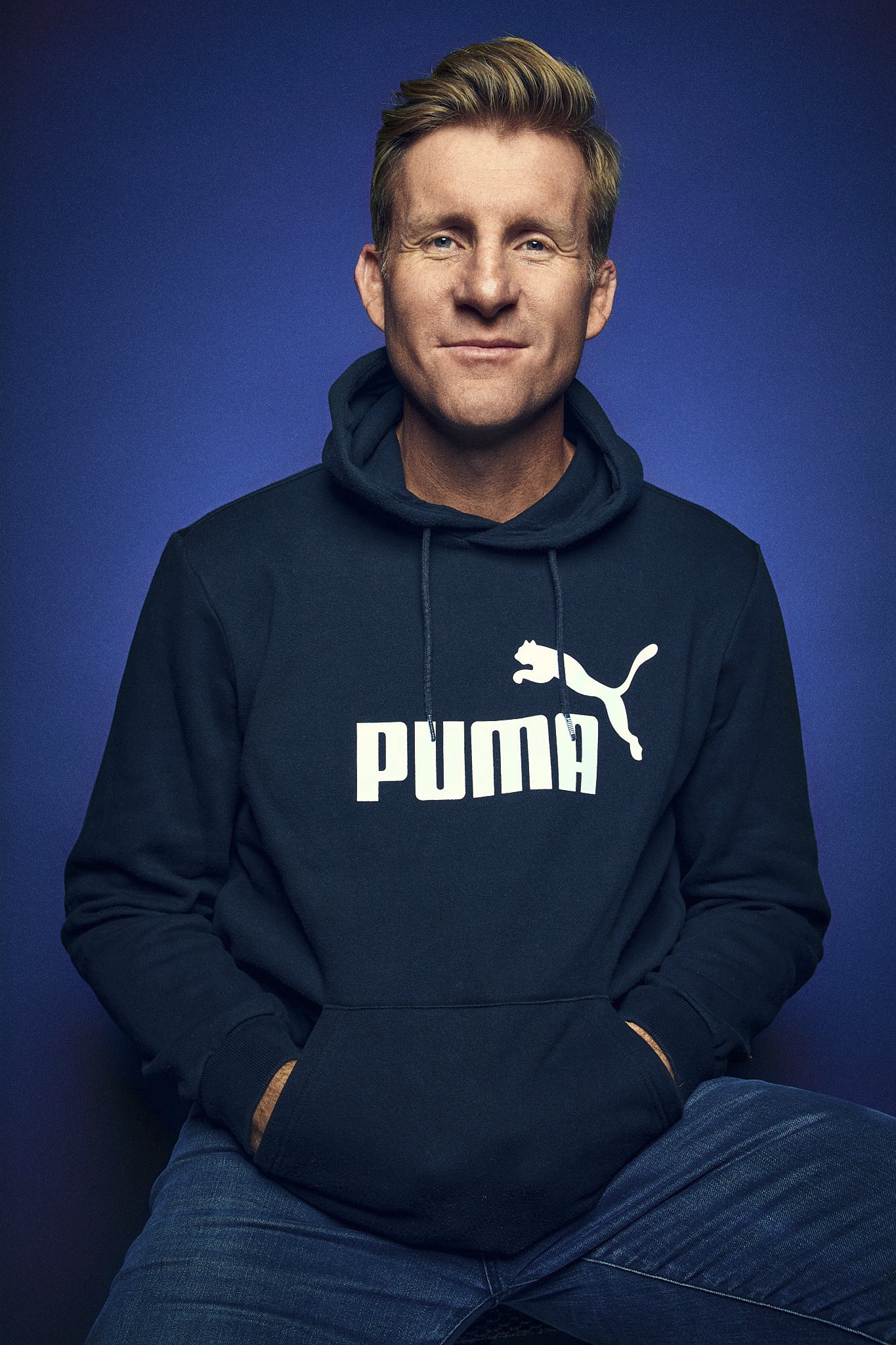 AJ Venter with short blonde hair wearing a dark Puma hoodie and blue jeans, sitting against a dark blue background.