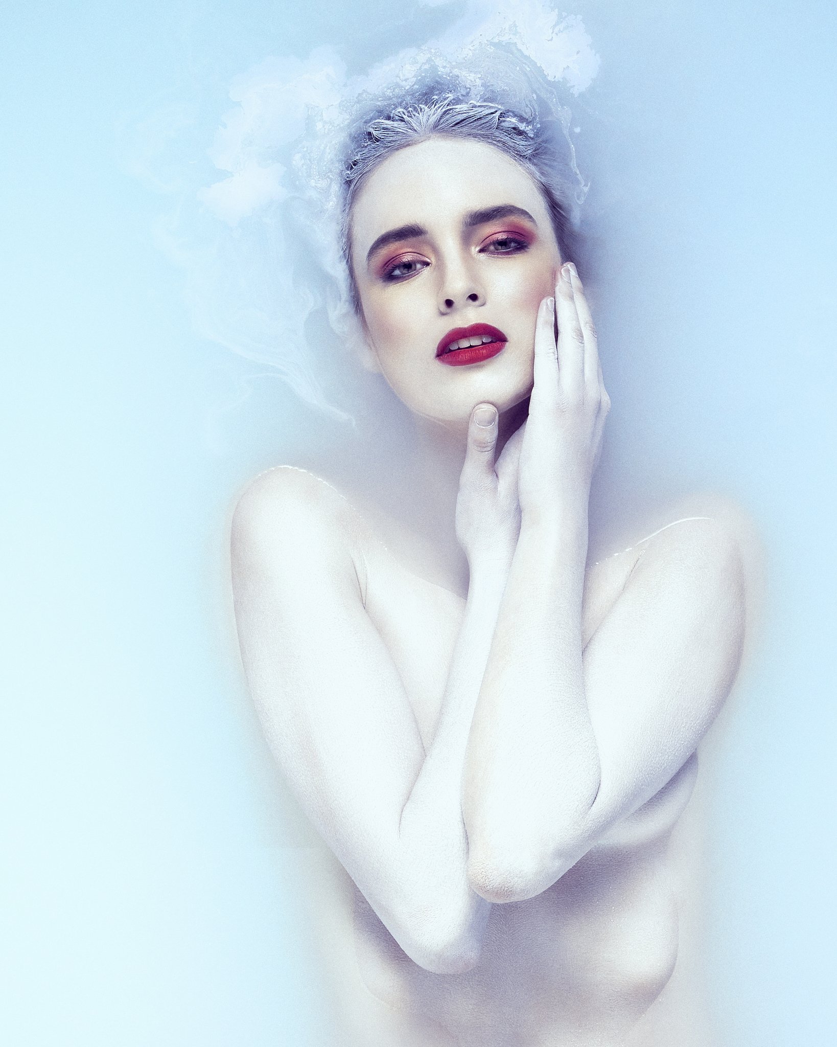 Close-up of a woman with fair skin and makeup, touching her face with her hand, floating in  light blue milky liquid.