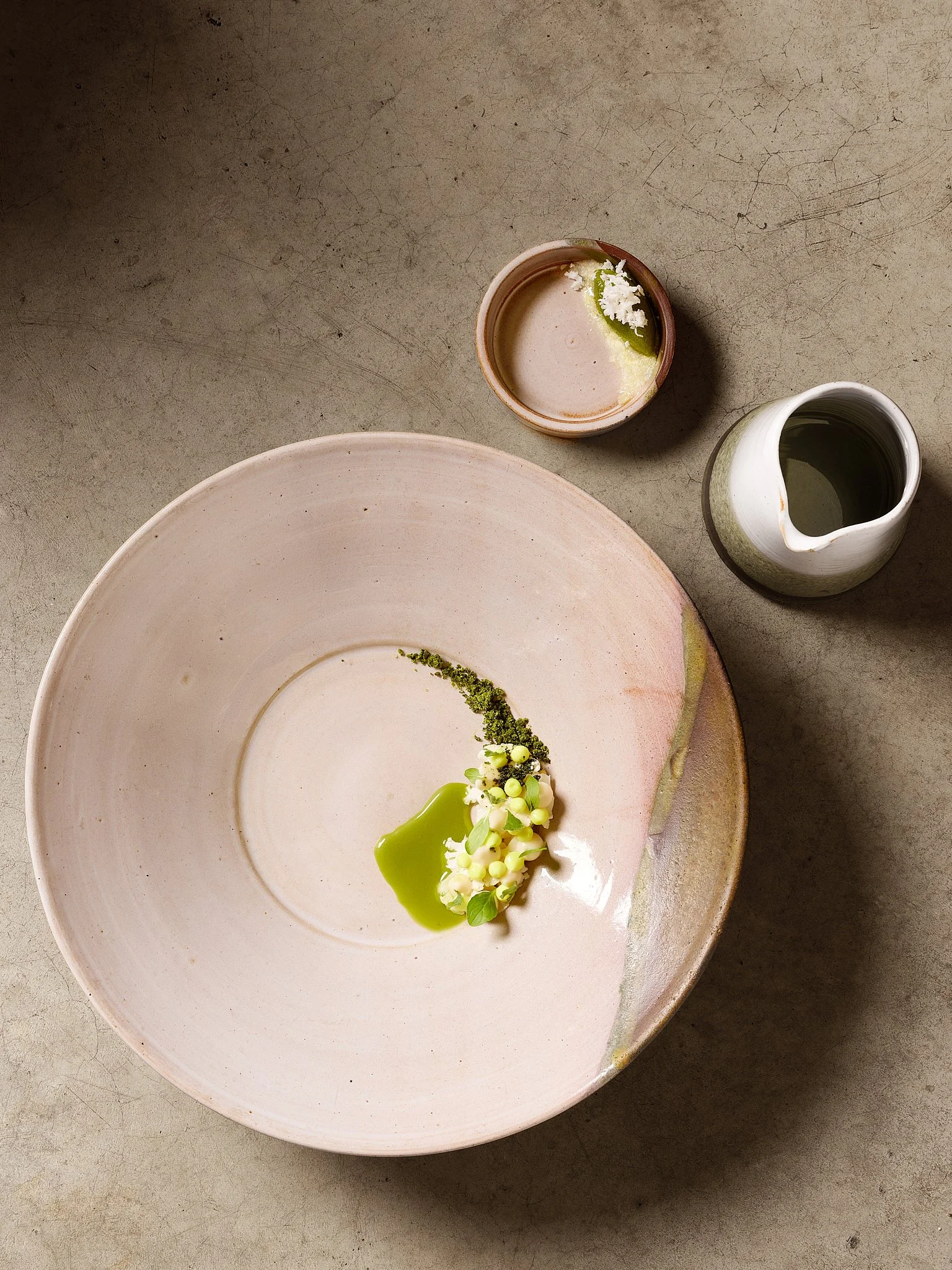 A large pink-rimmed plate with a green sauce, small white flowers, and green leaves, along with a small white pitcher filled with dark liquid, and a small ceramic cup with a lime wedge and white flower on top, on a gray textured surface.