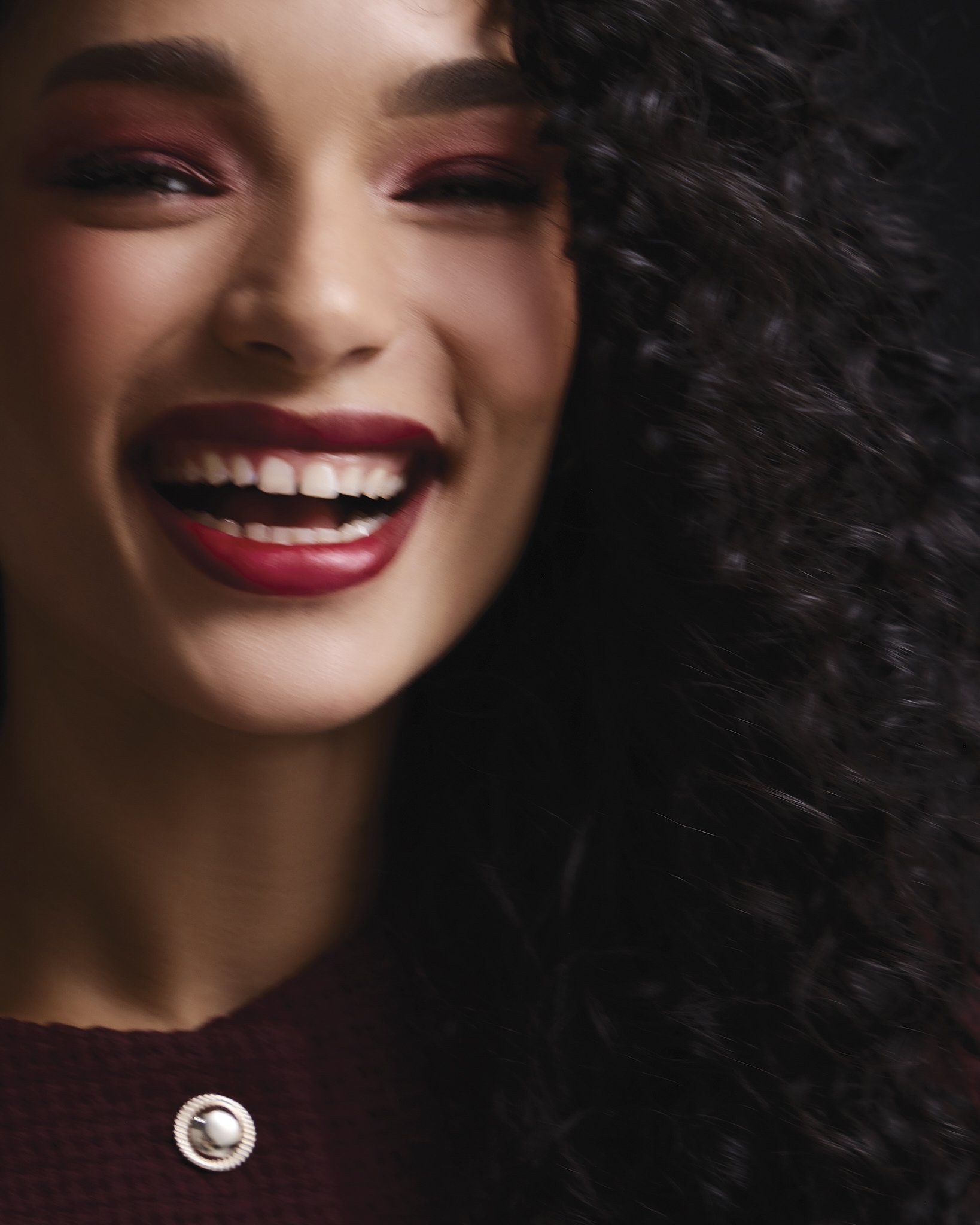Close-up of a woman with curly hair, smiling with red lipstick, wearing a maroon sweater with a white button.