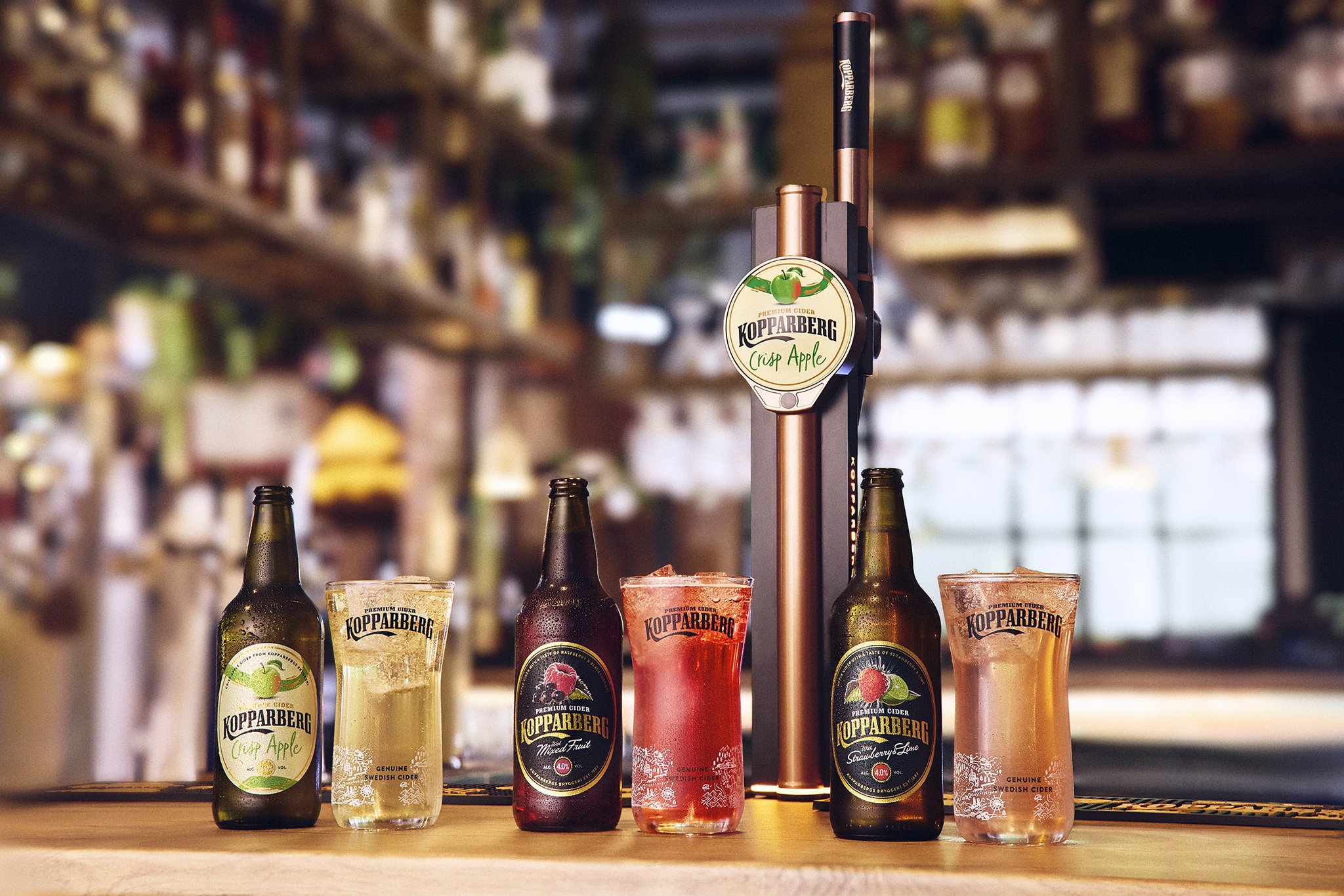 Various bottles and glasses of Kopparberg cider on a bar counter, with a blurred background of a bar or store shelves.