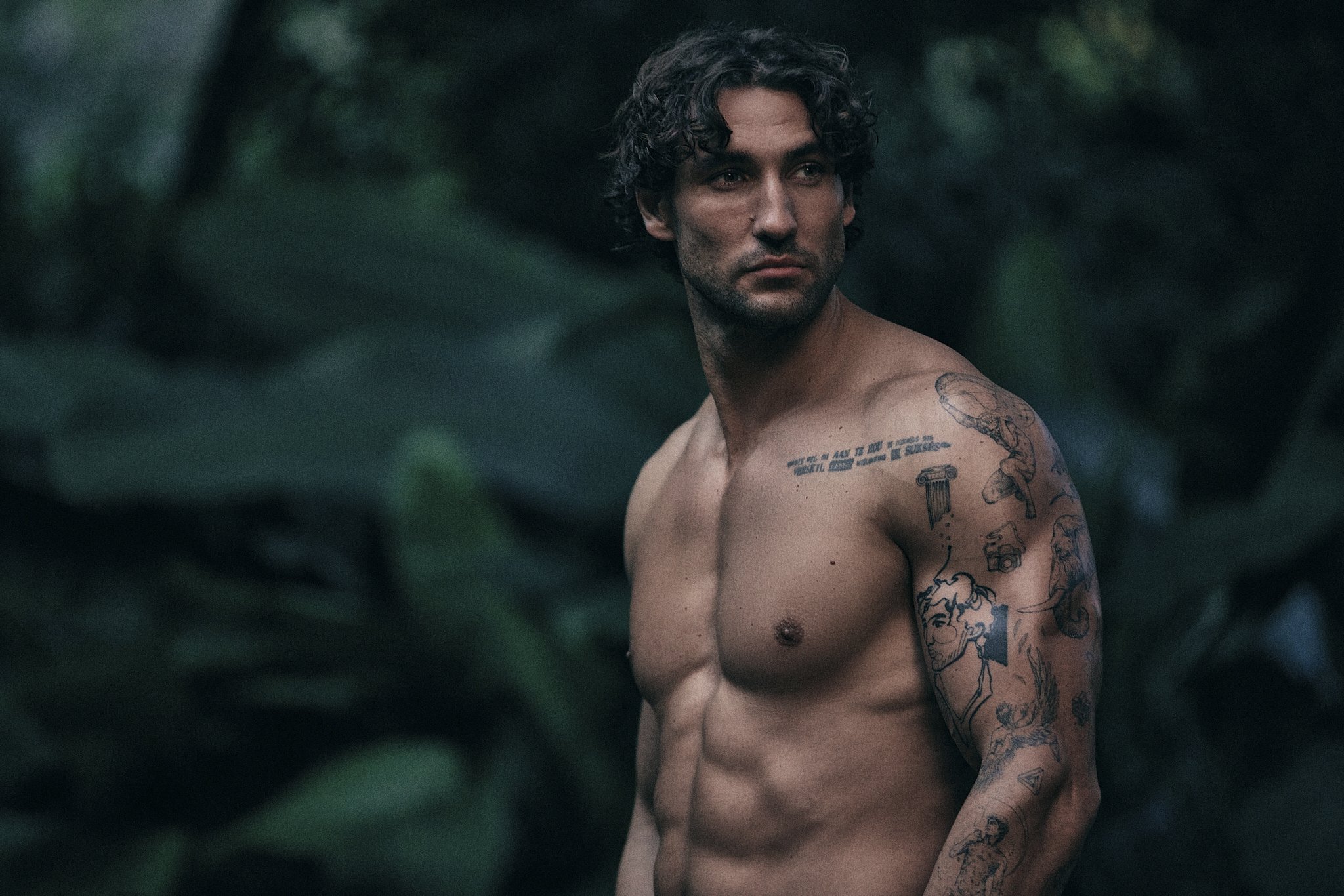 A shirtless man with curly dark hair and tattoos on his left arm and shoulder, looking over his shoulder in an outdoor setting with dark green foliage in the background.
