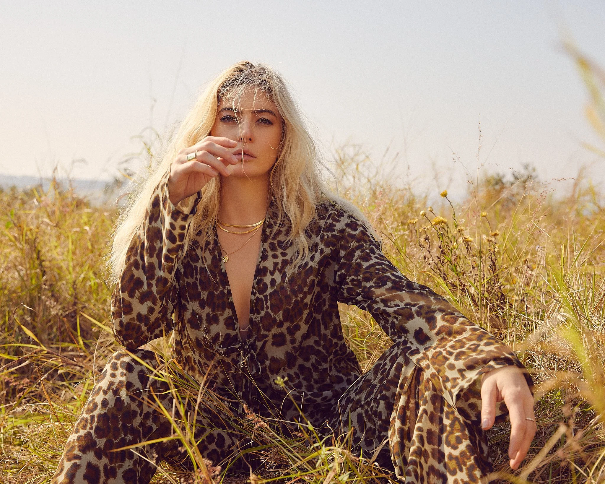 A beautiful blonde woman wearing leopard print clothing sitting in tall grass outdoors.
